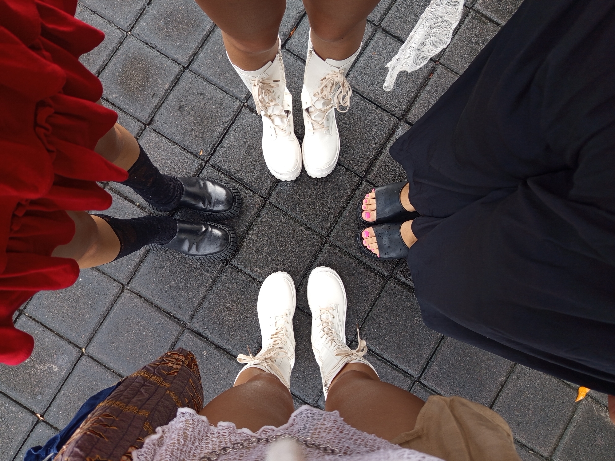 A high-angle shot captures the feet of four people standing on a grey brick-paved surface, likely a sidewalk or plaza. The composition features a circular arrangement of footwear.
A person to the left wears a short red skirt, black knee socks, and black leather shoes. The person above wears white boots with matching laces. To the right, a person wears a long black dress with black sandals, revealing pink toenails. The person below wears white boots with matching laces.
The location is North Kuta, Indonesia. The bricks are wet, suggesting recent rain. The lighting indicates daylight.