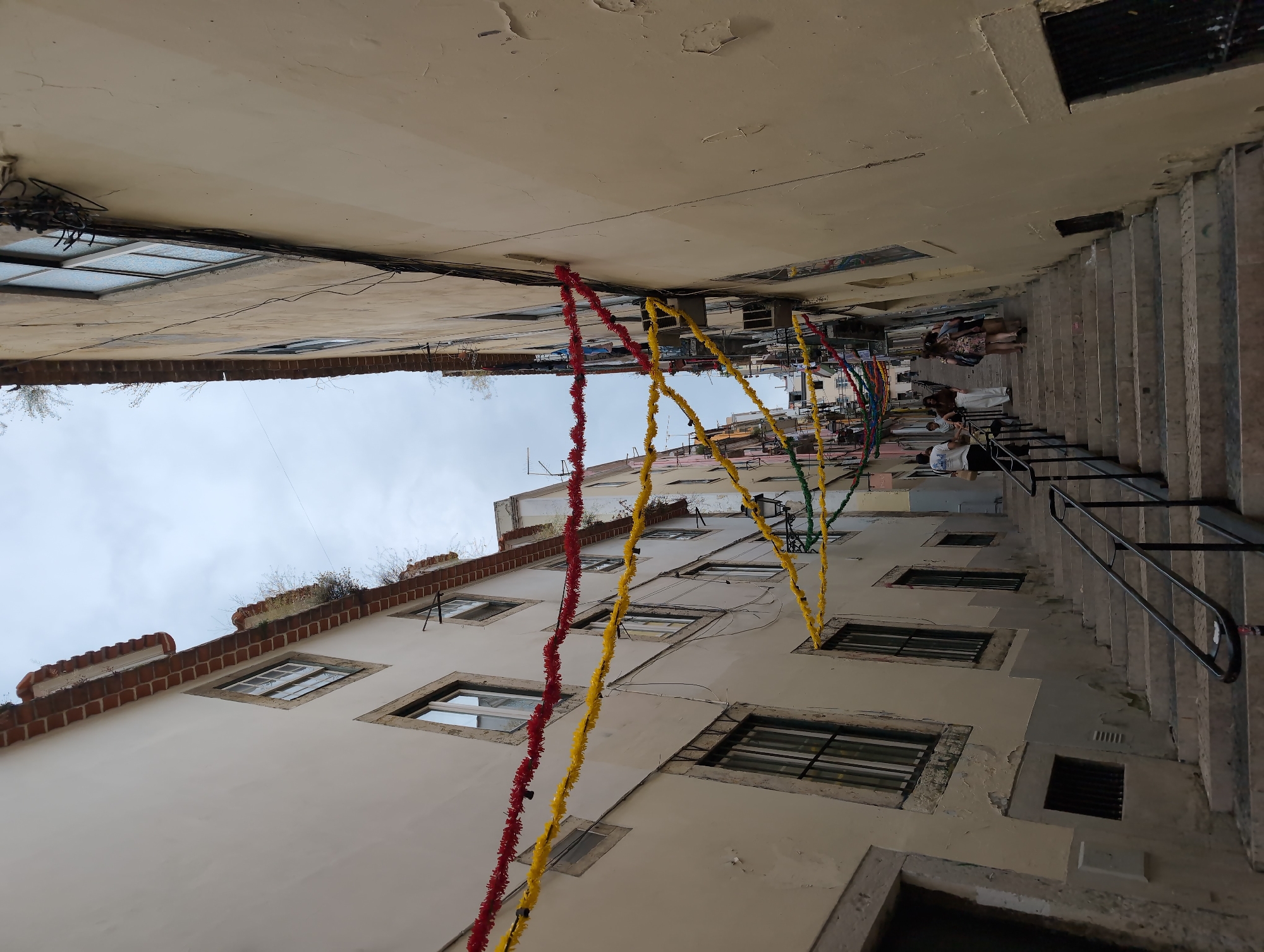 A low-angle shot looking up between buildings in an alleyway of Lisbon, Portugal. A set of steps lined with a black railing runs up the right side of the frame. People can be seen walking up the steps. Red, yellow, and green streamers hang overhead. The buildings are light beige. Windows are recessed into the walls. The sky is visible at the top of the frame.