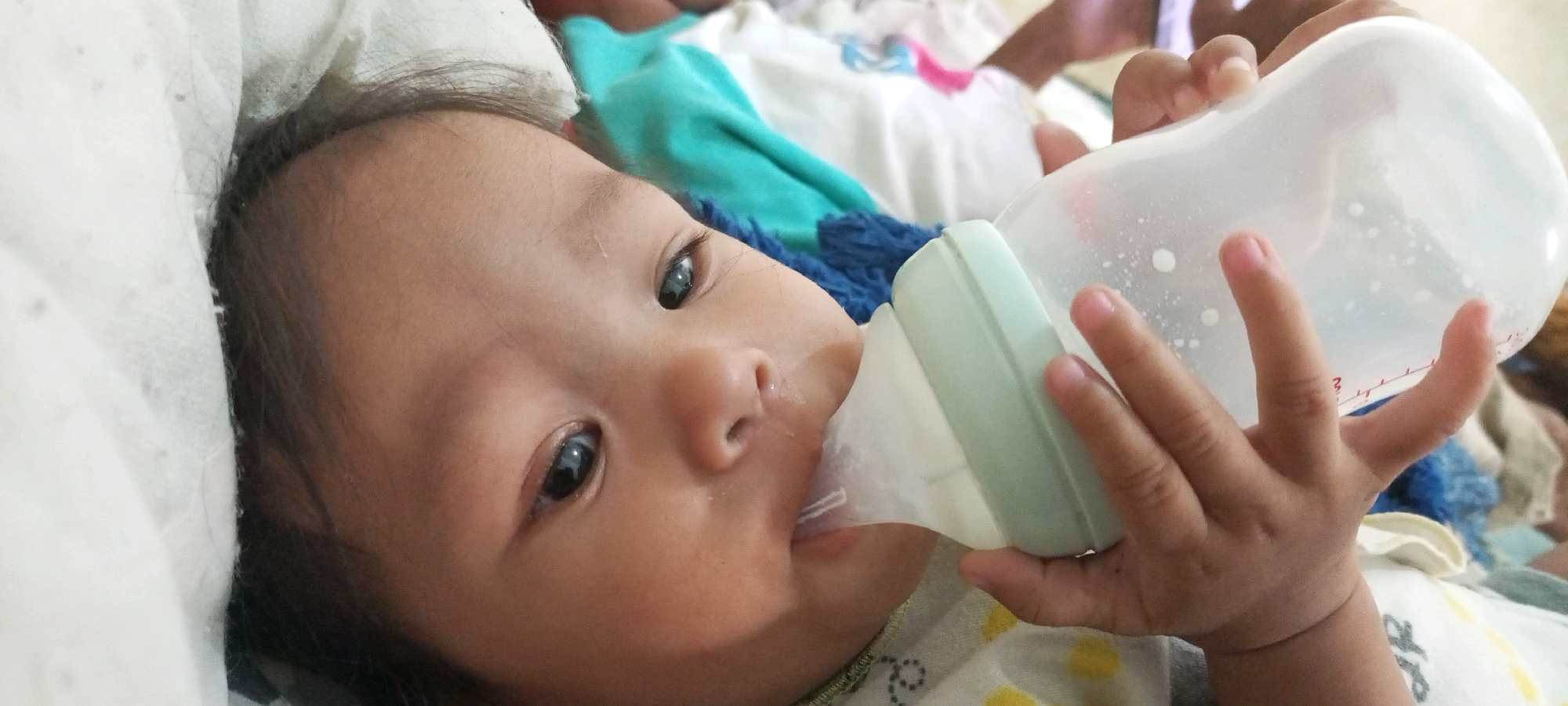 This is a close-up, horizontal, eye-level shot of a baby being fed from a bottle. The baby, likely of Asian descent, has dark hair, dark eyes, and a rounded face. The baby is lying down, propped up on a white surface that appears to be bedding or a blanket. The baby's mouth is open and engaged with the nipple of a transparent plastic baby bottle, which is held by an adult's hands. The bottle contains a white liquid, possibly milk, with small droplets visible inside. A pale green collar on the bottle connects the nipple to the main body. The background is softly blurred, showing glimpses of colorful fabric, including teal and white, suggesting a domestic setting. The lighting is natural and appears to be from daytime. The overall mood is intimate and caring, focused on the feeding moment. The visible text on the bottle is "AR".