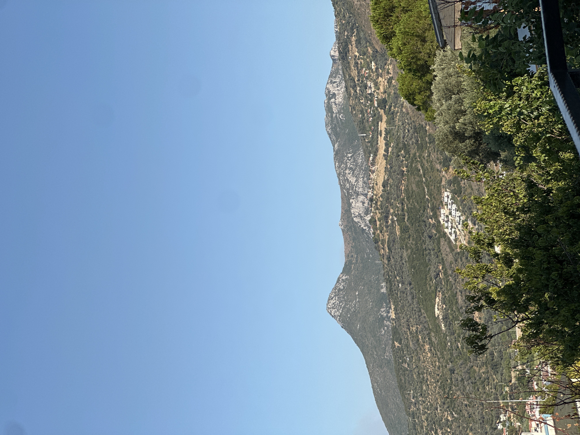 This is a photograph taken from a high vantage point, likely a balcony or elevated structure, overlooking a hilly landscape near the city of Karaburun, Turkey. The image is dominated by a clear, light blue sky, which occupies the top half. Below the sky, a range of mountains stretches across the horizon. The mountains are covered in dense green vegetation, with patches of lighter brown and rocky outcrops visible on their slopes.
In the mid-ground, nestled within the foothills of the mountains, there is a scattering of white buildings, indicative of a settlement or village. A slender white minaret rises above the buildings, suggesting the presence of a mosque. The terrain is uneven, with terraced slopes and winding paths or roads visible. Olive trees and other Mediterranean flora are abundant, creating a textured, green tapestry across the hillsides.
In the foreground, a dense collection of green foliage, including leafy trees and branches, partially obscures the view of the landscape. A dark metal railing is visible at the very bottom of the frame, further framing the scene from the perspective of the observer. The overall impression is one of a rural or semi-rural setting in a hilly coastal region under a bright, clear sky.