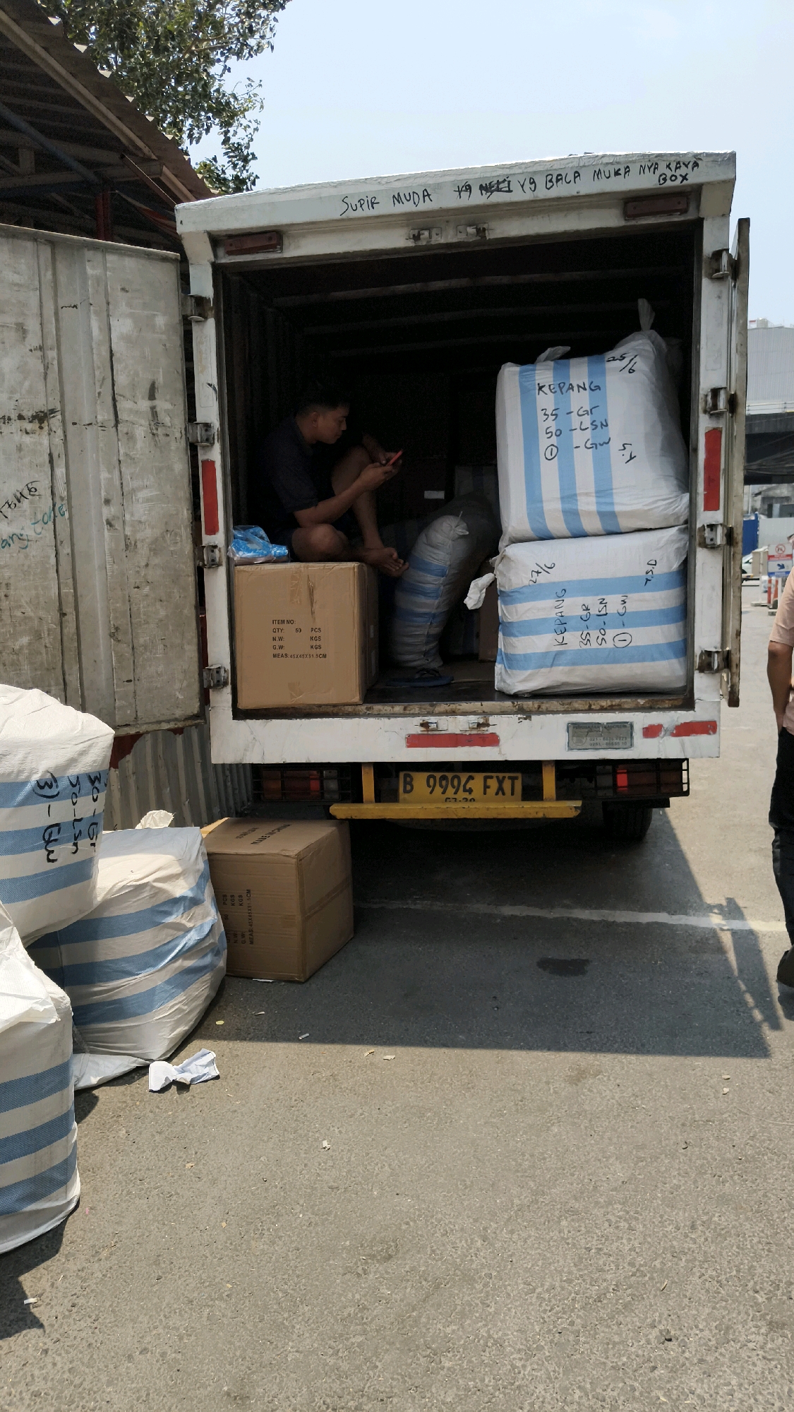 A man sits inside the open rear of a white delivery truck, looking at his phone. He is wearing a dark shirt and shorts, and his legs are crossed. The truck is filled with large white sacks with blue stripes, and a cardboard box. The sacks have writing on them in blue marker. The truck's license plate is B 9994 FXT. Outside the truck, more sacks are visible. The scene appears to be in an urban or industrial area, with a building and trees in the background. The ground is paved. The overall impression is of a logistics or delivery operation in progress.