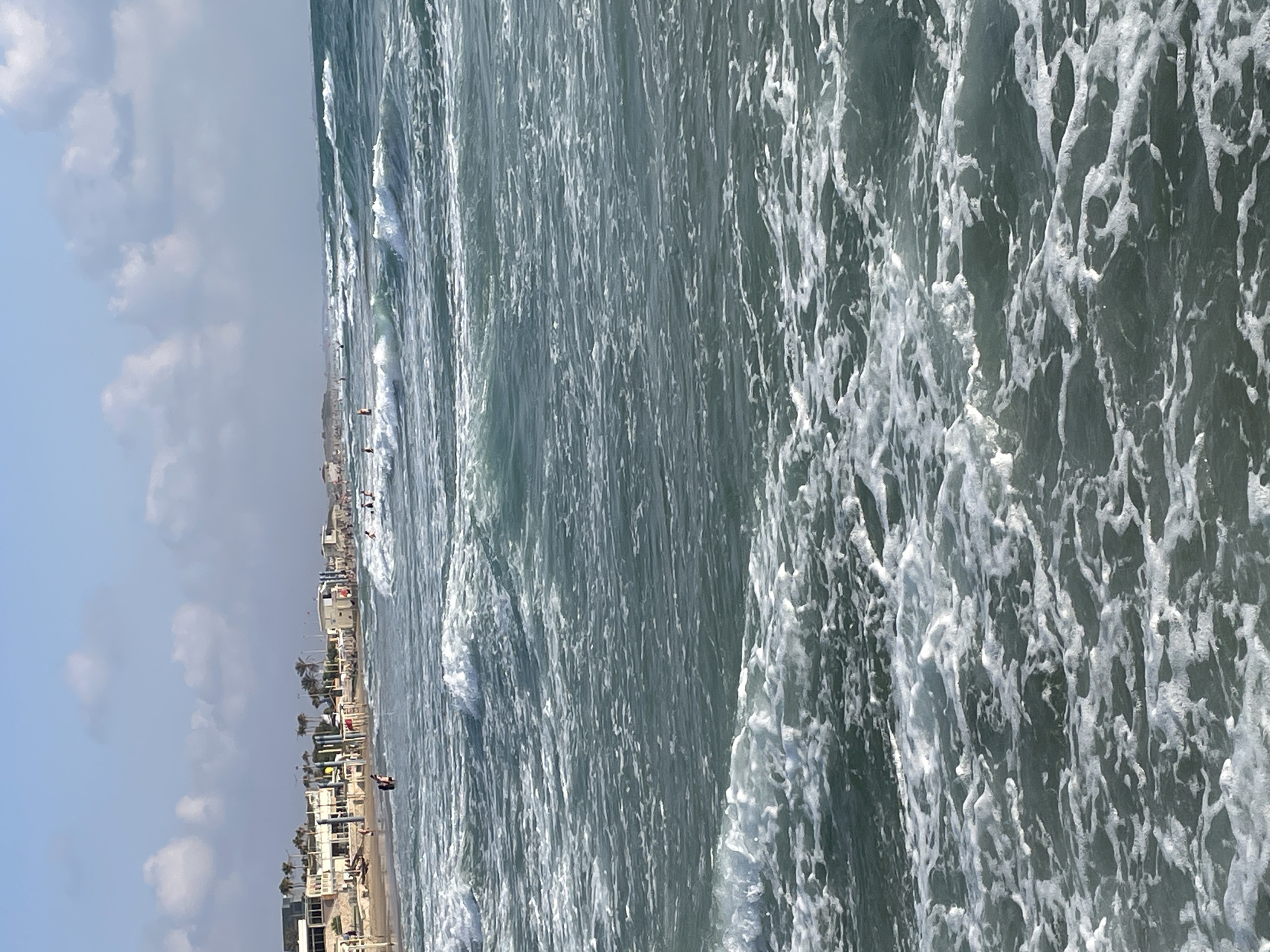 The image shows a view of the sea with waves crashing on the shore. In the background, there is a beach with people and buildings. The sky is blue with some clouds. The scene appears to be a busy beach on a sunny day. This location is likely one of the beaches in Haifa, Israel, given the architecture visible in the background, which is typical of Israeli coastal towns.
