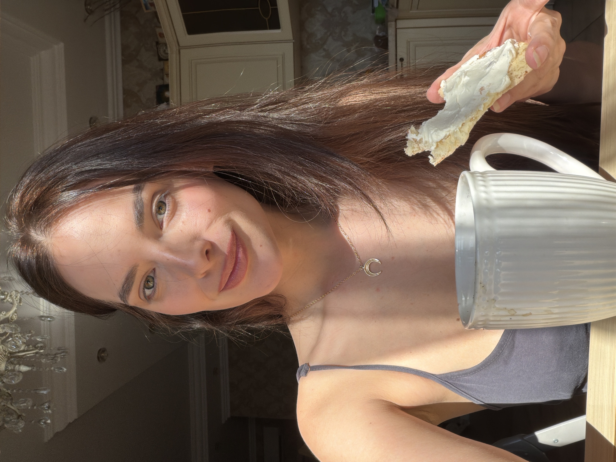 In an indoor setting, likely a kitchen, a woman with long brown hair and green eyes is captured in a medium shot, holding a piece of bread with cream cheese and a white mug. Sunlight streams in from the left, casting shadows and highlighting her features. She is wearing a dark gray tank top and a delicate gold necklace with a crescent moon pendant. The background reveals kitchen cabinets and a chandelier, suggesting a home environment. The woman is looking at the camera with a gentle smile, and the overall mood is relaxed and natural.