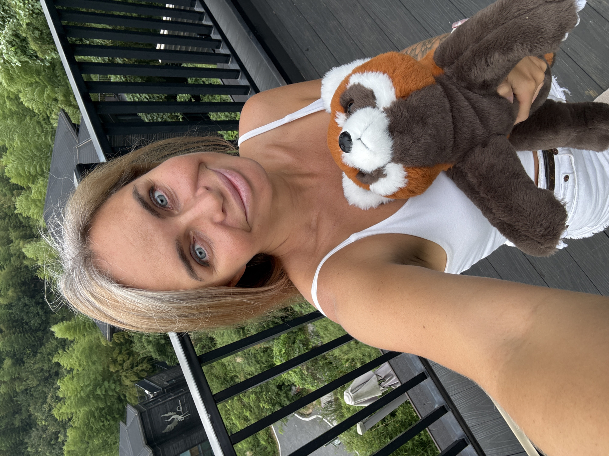 A woman with blonde hair and blue eyes is taking a selfie on a wooden balcony. She is holding a stuffed red panda. In the background, there are trees and buildings. The location is Wuxing District, China.