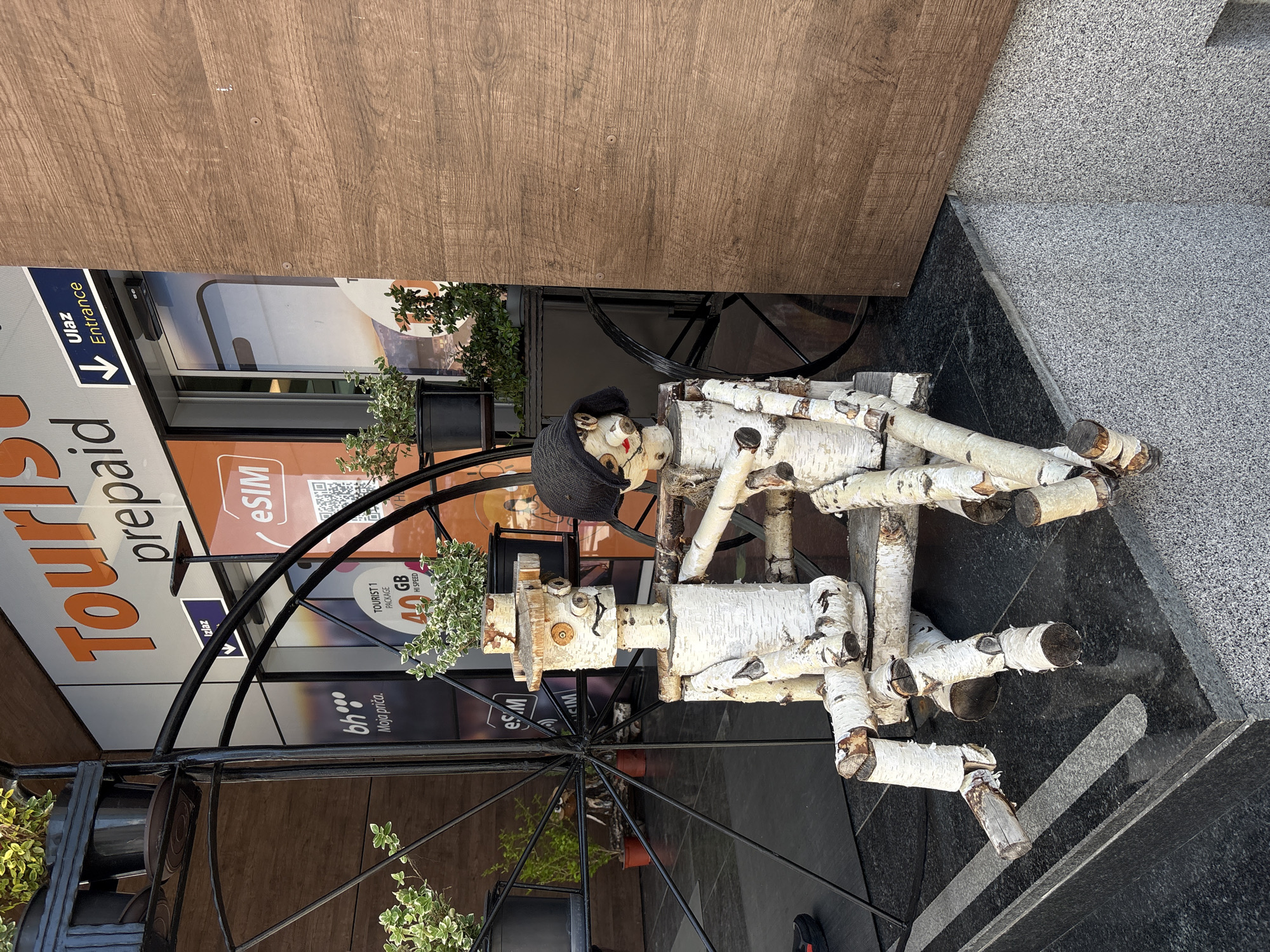 The image shows a wooden sculpture of a person made from birch logs, sitting on a black metal bench with flower pots hanging on the sides, positioned outside a storefront in Sarajevo, Bosnia and Herzegovina. The storefront has a sign visible that reads "Tourist prepaid eSim" and includes details about tourist packages with data. The floor is dark grey stone, and a wooden wall is in the background.