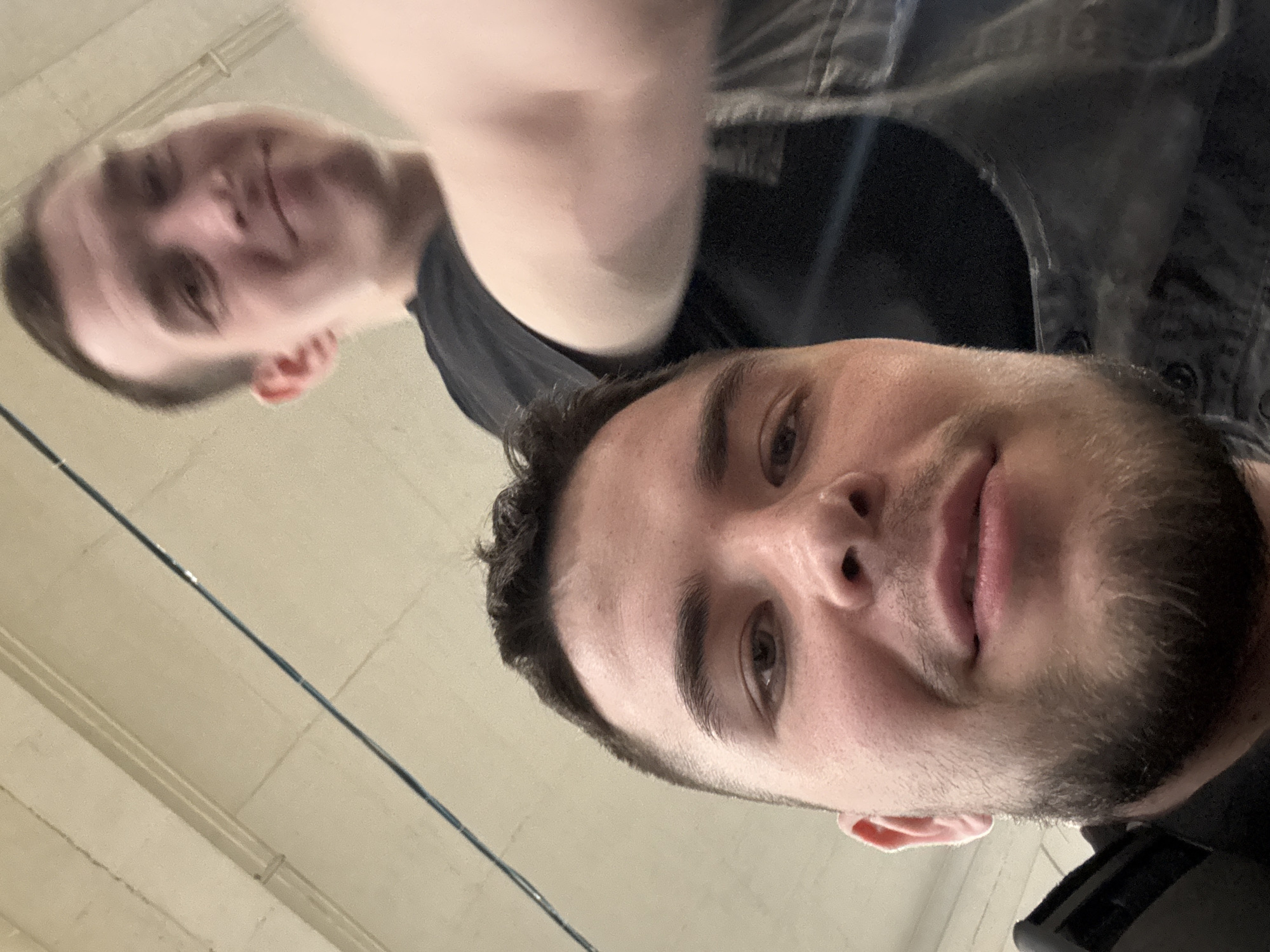 Two males are captured in a low-angle, close-up selfie photograph in an indoor setting in Dikaya Gar', Russia. The individual in the foreground occupies the majority of the lower half of the frame, presented sharply in focus. He has short, dark hair and a neatly trimmed dark beard and mustache, with visible stubble on his cheeks and chin. His eyes are dark, and he has a subtle, closed-mouth smile. He is wearing a dark-colored garment, possibly a shirt or jacket, with a black strap or element visible on his left shoulder.

Behind and slightly to the right of the foreground individual, a second male is visible from the chest up, positioned higher in the frame and appearing soft-focused. He has short, dark hair and a light complexion, with a wider, open-mouth smile. He is wearing a plain black t-shirt beneath a dark denim garment, possibly overalls or a jacket, the shoulder strap of which is visible on his right side. His right forearm is blurred and extends into the right mid-frame, appearing to be lifted towards the camera.

The background consists of a light-colored ceiling with visible seams or paneling, indicative of an industrial or institutional interior. A thin, dark cable or pipe runs diagonally across the upper left portion of the ceiling. The overall lighting is even, suggesting an adequately lit indoor environment.