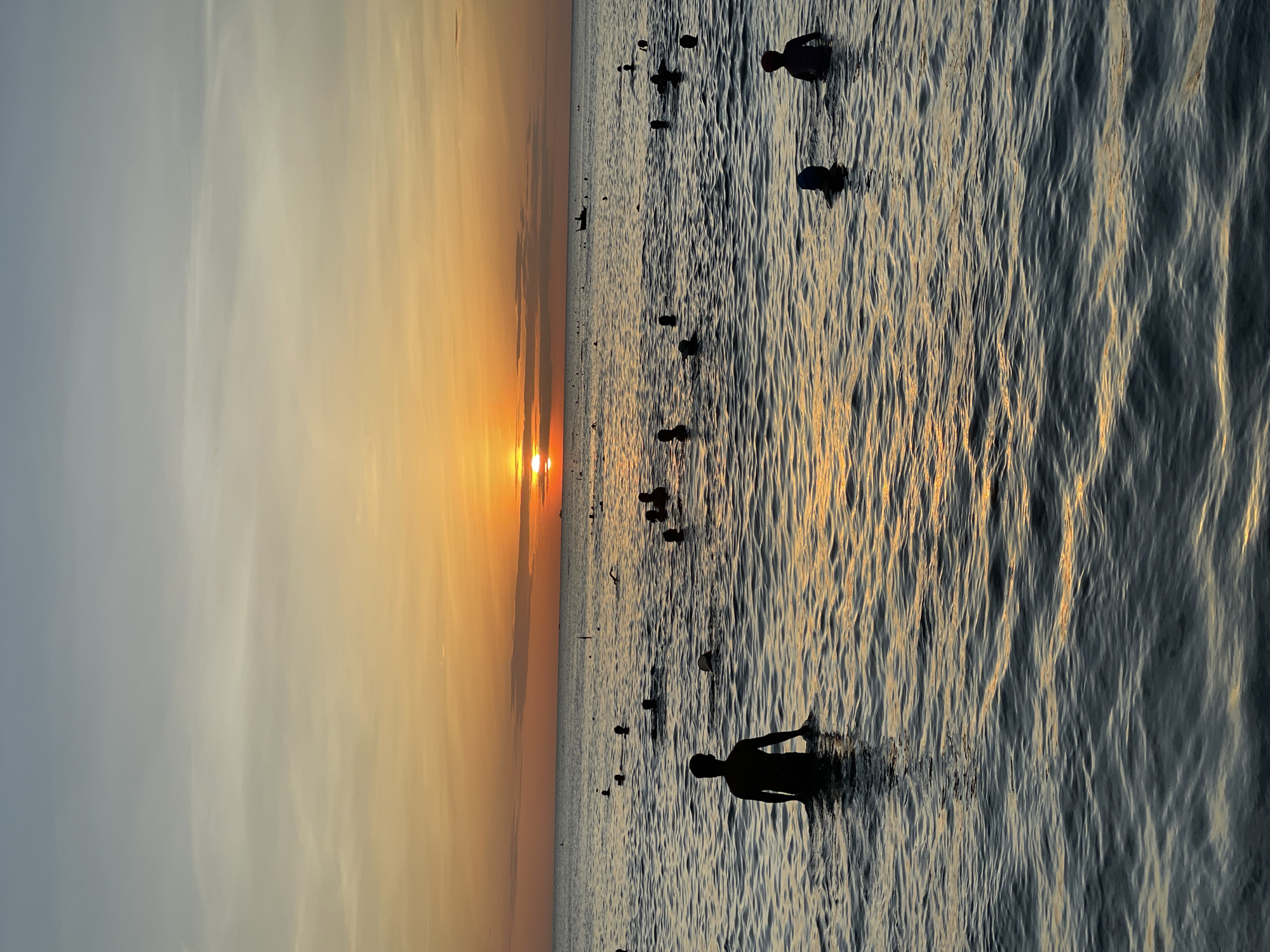 The image depicts a sunset over the ocean, with numerous people swimming and wading in the water. The sun is setting on the horizon, casting an orange glow across the sky and reflecting on the rippling surface of the sea. Silhouettes of people are visible in the water, some standing, some swimming, and some with their heads above the surface. In the foreground, a person stands in the water, facing away from the camera towards the sunset. In the distance, a small boat can be seen on the water. The scene suggests a popular beach destination, likely in the city of Đà Nẵng, Vietnam, known for its beautiful coastline and vibrant sunset views. The water is calm with gentle waves, and the overall atmosphere appears relaxed and serene.