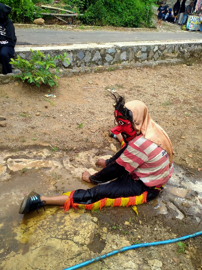 An individual is depicted seated on muddy ground in Gebyog, Indonesia. The person wears a red mask with an elongated nose, white eyes, and spiky black hair, topped by a light beige hood or scarf. They are dressed in a red and white striped t-shirt, black trousers, and red and yellow fringed leg coverings around the ankles. The left foot is shod in a black athletic shoe with white stripes, while the right foot is bare and partially immersed in a puddle. Both hands are bare and rest on the muddy surface. The immediate foreground consists of dirt mixed with standing water and exposed cracked ground. A blue hose or pipe runs diagonally across the lower right. Behind the seated individual, a dirt area leads to a stone retaining wall. Beyond the wall is a dark paved road, bordered by dense green foliage. In the far background, multiple figures are visible on the road, some gathered near a vendor stall with a colorful banner. Another person in a black hoodie is seated on the dirt in the far left background.