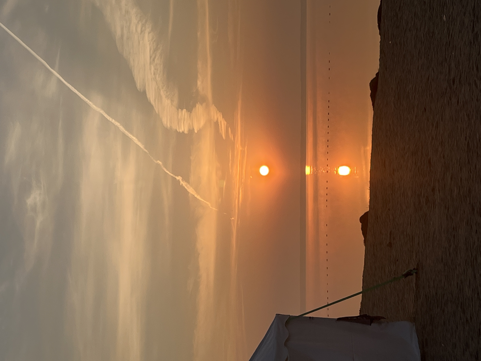 A sunrise casts a warm, orange glow over a tranquil body of water, reflecting the sun's brilliant orb. The scene is set on a sandy beach, with a white tent visible on the left, its green support pole anchored to the ground. In the distance, a faint line of buoys stretches across the water. The sky is a canvas of soft clouds, streaked with a long, white contrail from an aircraft. The overall impression is one of peaceful solitude in Surakovo, Russia.