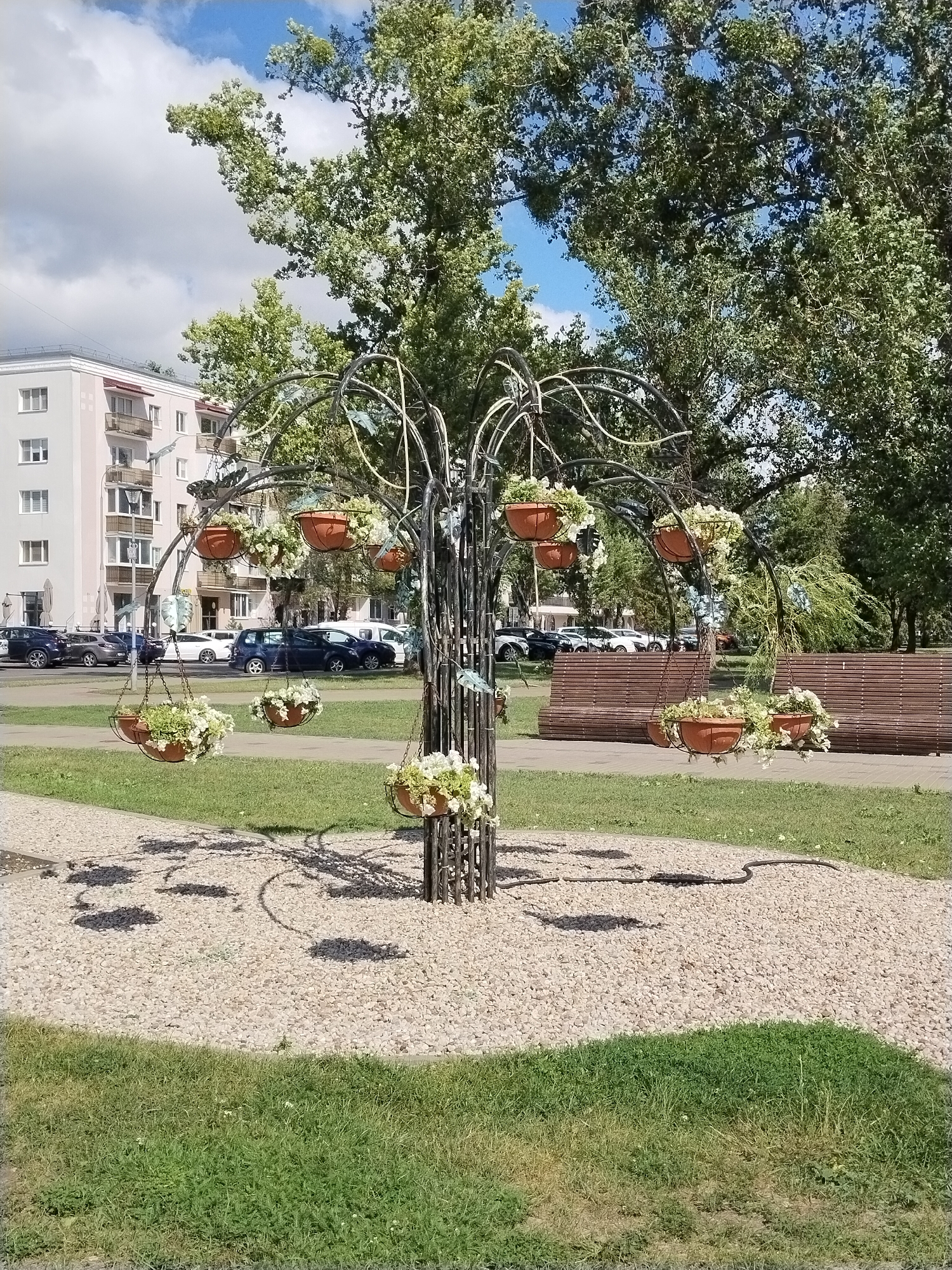 This is a photograph taken on a sunny day in the city of Brest, Belarus, specifically at the intersection of Maskouskaia Street and Kavalioff Street, in front of apartment building number 44. The central focus of the image is a decorative metal tree sculpture with numerous hanging planters filled with white flowers. These planters are suspended from curved metal branches, creating a whimsical display. The sculpture is situated on a bed of light-colored gravel, which contrasts with the surrounding green grass. In the background, a light-colored apartment building with balconies and windows is visible. Further in the distance, cars are parked along a street, and park benches made of wooden slats are arranged in a grassy area. Tall trees with lush green foliage dominate the upper portion of the image, with patches of blue sky and white clouds visible through their branches. The overall scene depicts an urban park or public space adorned with artistic landscaping.