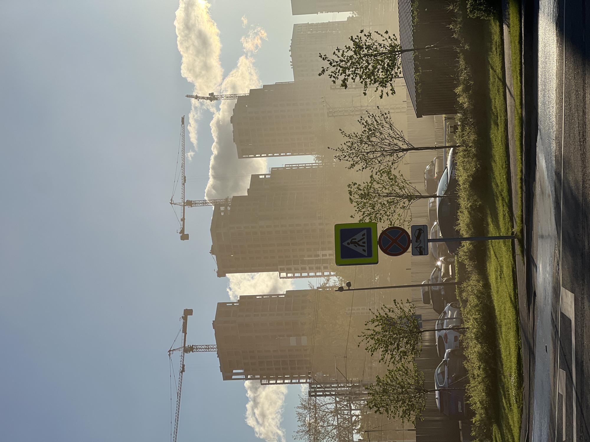 The image depicts a hazy, sunlit scene of a construction site in Khimki, Russia. In the background, several unfinished high-rise buildings under construction are visible, with cranes towering over them. The sun, positioned to the right of the frame, casts a warm glow and creates lens flare, contributing to the atmospheric haze. In the foreground, a street with a crosswalk is visible, with several cars parked along the side. Green grass and sparse trees line the area, suggesting a developed urban environment. Traffic signs, including a pedestrian crossing sign and a "no stopping" sign, are present near the road. The overall impression is one of ongoing development and urban expansion under a bright, possibly dusty or humid, sky.