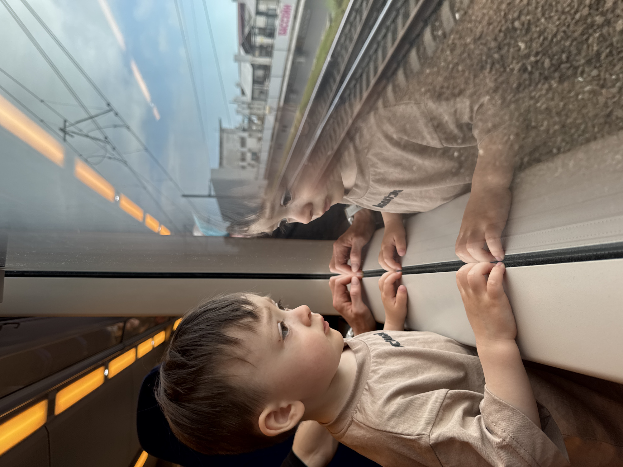 A young child with brown hair and wearing a tan t-shirt is looking out of a train window. The child's hands, along with an adult's hands, are touching the window frame. The reflection in the window shows the child's face and shirt, along with the passing landscape. The image seems to be taken from inside the train, with lights visible along the side of the train car. Outside the window, railway tracks, buildings, and other structures are visible. In particular, the building reads "SOCHIN." The image is taken in Sochi, Russia.