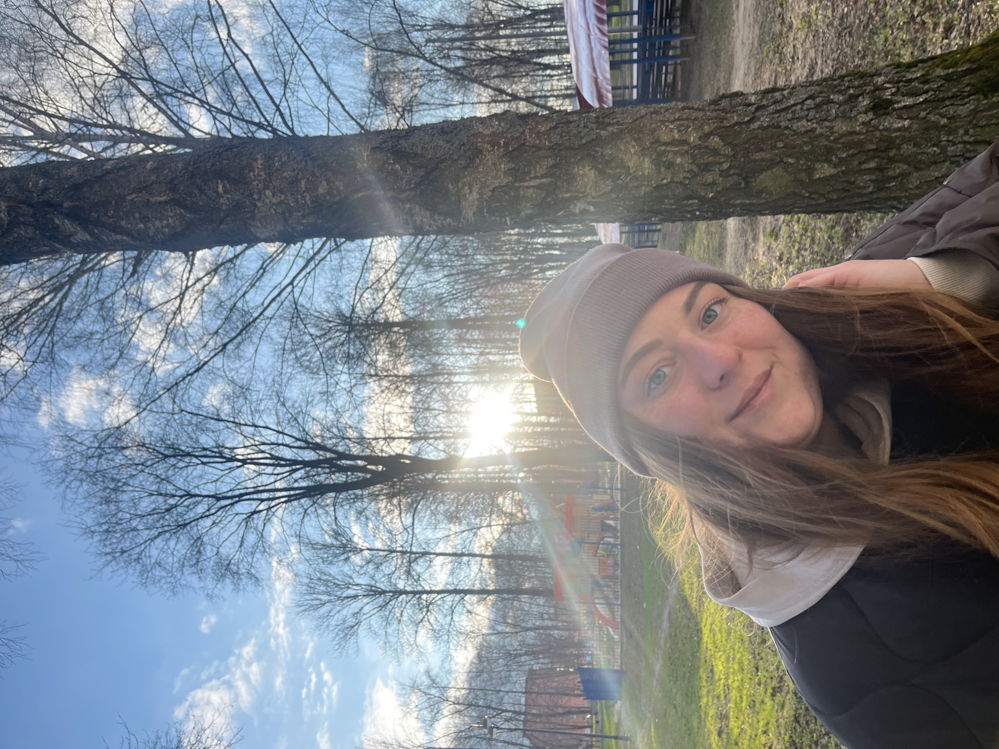 A selfie of a young woman with long brown hair wearing a grey beanie and a dark jacket, standing outdoors in a park in Klin, Russia. Trees with bare branches are visible in the background, as well as a patch of green grass. The sky is blue with some clouds. The sun is shining and creating a bright glare in the image. A fence and playground equipment are visible in the background, suggesting a park or recreational area.