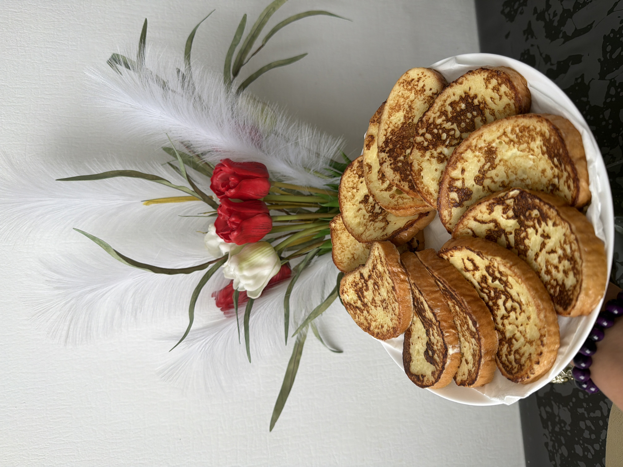Here's a description of the image:
The image shows a plate of what appears to be French toast, arranged in a semi-circle. Next to the plate is a bouquet of red and white tulips, interspersed with feathery white plumes and some green foliage. The background is a mix of white and a darker, marbled gray surface. A part of a person's arm can be seen at the right edge of the photo, with a purple beaded bracelet on the wrist. The image doesn't provide any clues about the location or city, so it is Unknown, Unknown.