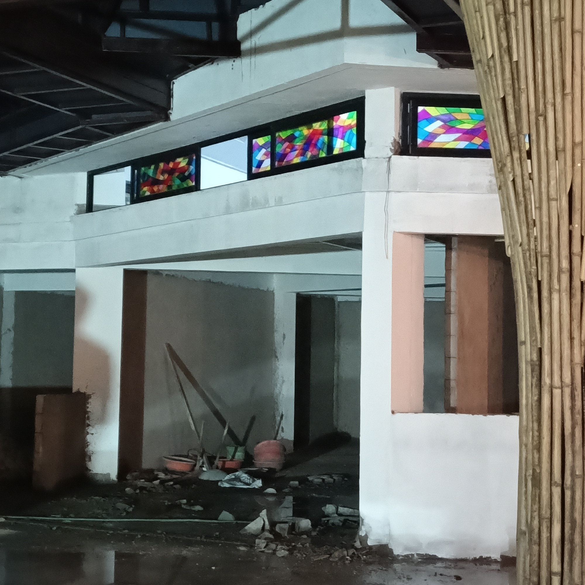 The image shows an unfinished building under construction. The structure is primarily made of concrete, with several open doorways and unfinished walls. Stained-glass windows are set into the upper part of the building, featuring colorful geometric patterns. Construction tools, including shovels and buckets, are scattered on the ground, amidst debris and rubble. A bamboo screen is partially visible in the foreground on the right side of the image. The building appears to be located outdoors, possibly in the city of Panaji, India, although there are no specific landmarks or details to confirm the location. There are no people visible in the image.