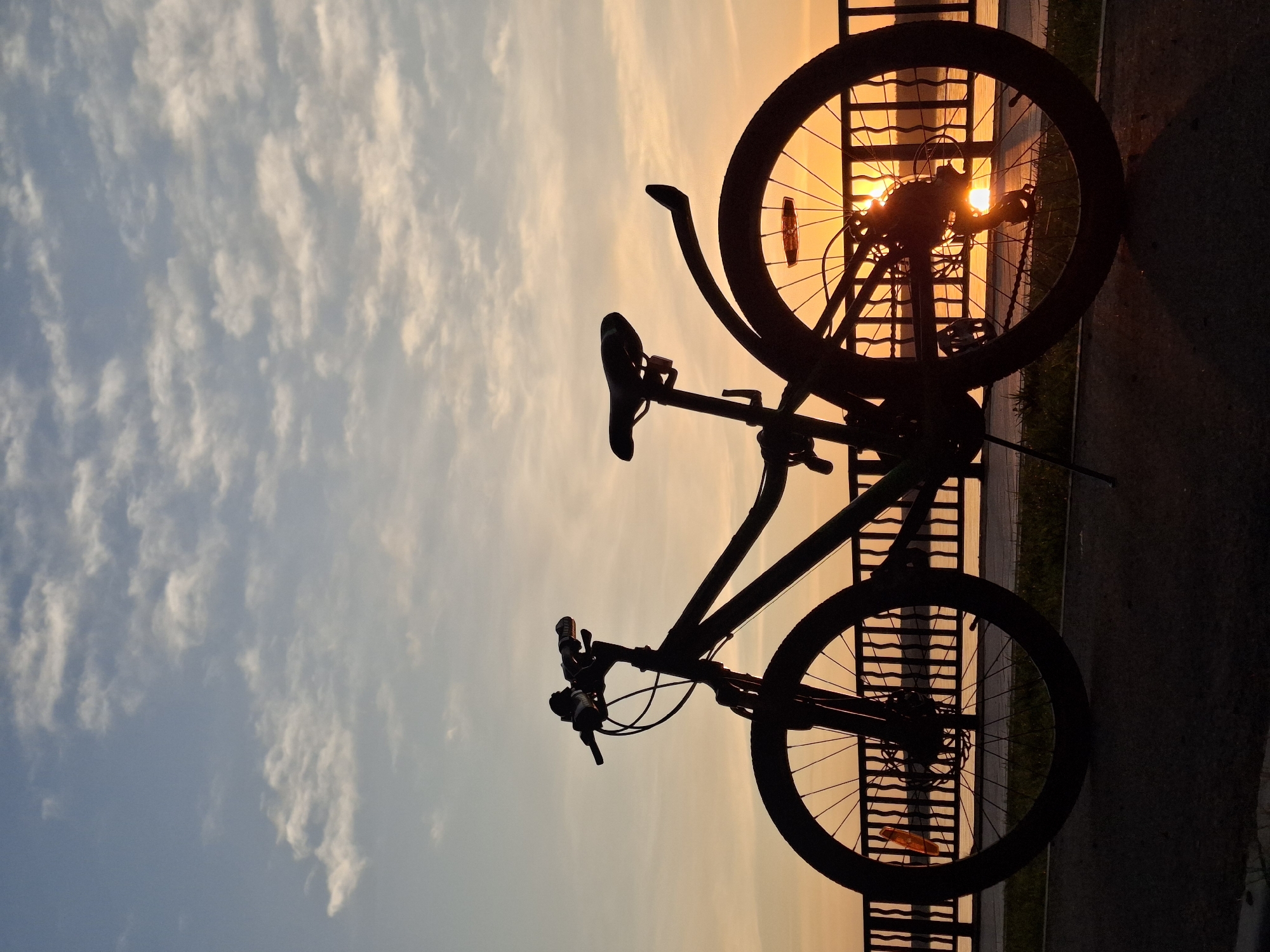 A silhouette of a bicycle stands against a sunset in Chaikovsky, Russia. The bike, positioned in front of a barrier along a walkway or path, is the central subject. The backdrop features a sky gradient of light blue to orange, with scattered clouds. The fence is low, possibly overlooking a body of water or an open area. The sun's rays peek through the bicycle wheel, creating a starburst effect.