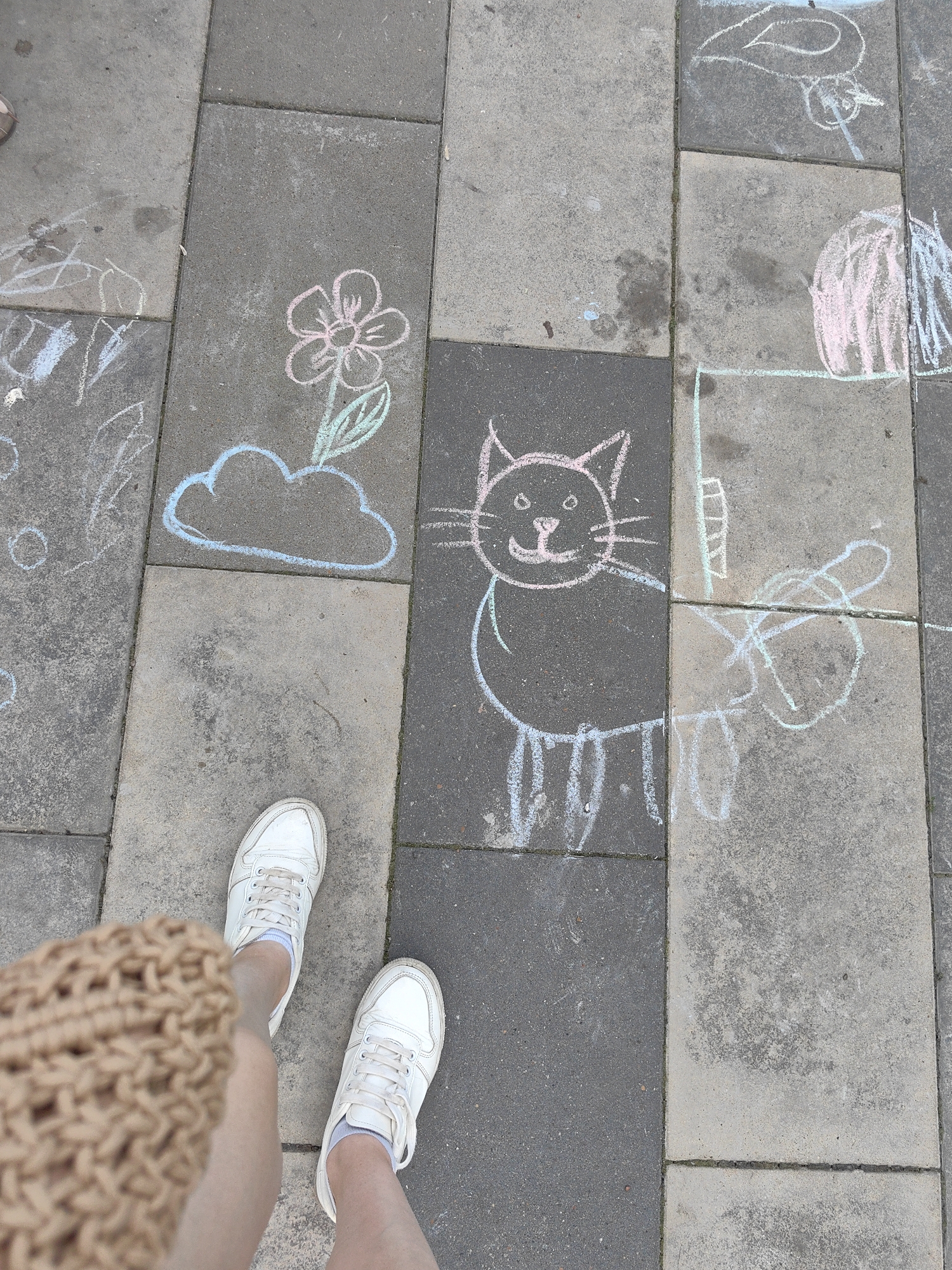 The image shows a close-up, overhead view of two feet wearing white sneakers and grey socks, standing on a paved walkway. The walkway is made of rectangular grey pavers. In the background, chalk drawings are visible on the pavers, including a pink and blue flower with a cloud, and a pink cat with a green tail and blue legs. The chalk drawings appear to be child-like in style. Some of the chalk drawings are faded or partially obscured by water stains on the pavers. The overall scene suggests an outdoor environment, possibly a park or sidewalk in a city. The specific location of Ufa, Russia is not evident from the image itself, as there are no distinguishing landmarks or text.