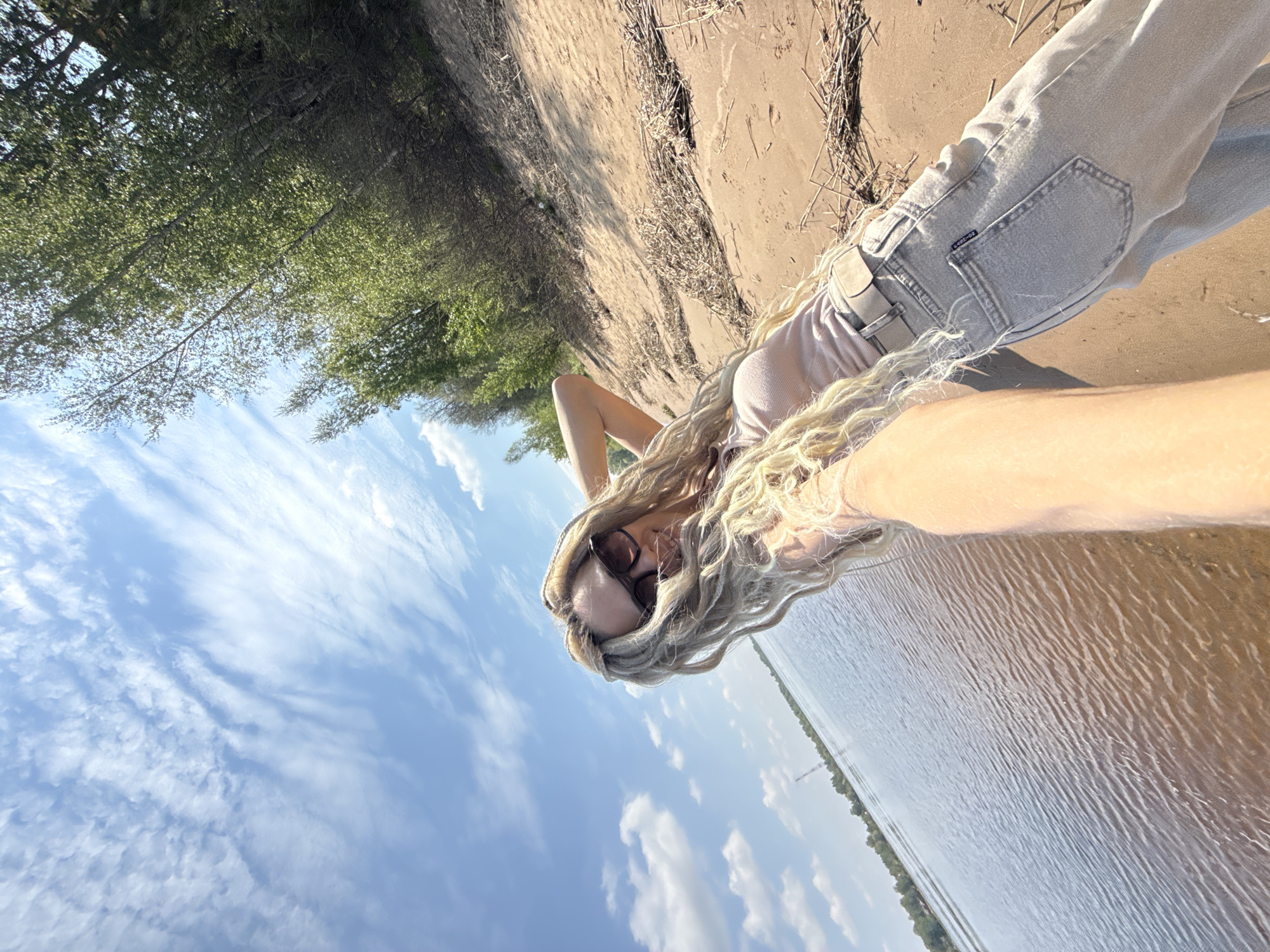 A person lies on their side on a sandy beach in Rusanovo, Russia, with their head towards the blue, partly cloudy sky and their body angled towards a body of water. The person is wearing a light-colored top and jeans, and sunglasses. They have long, blonde hair that obscures part of their face. The environment includes trees and vegetation along the shore and sand along the edge of the water.