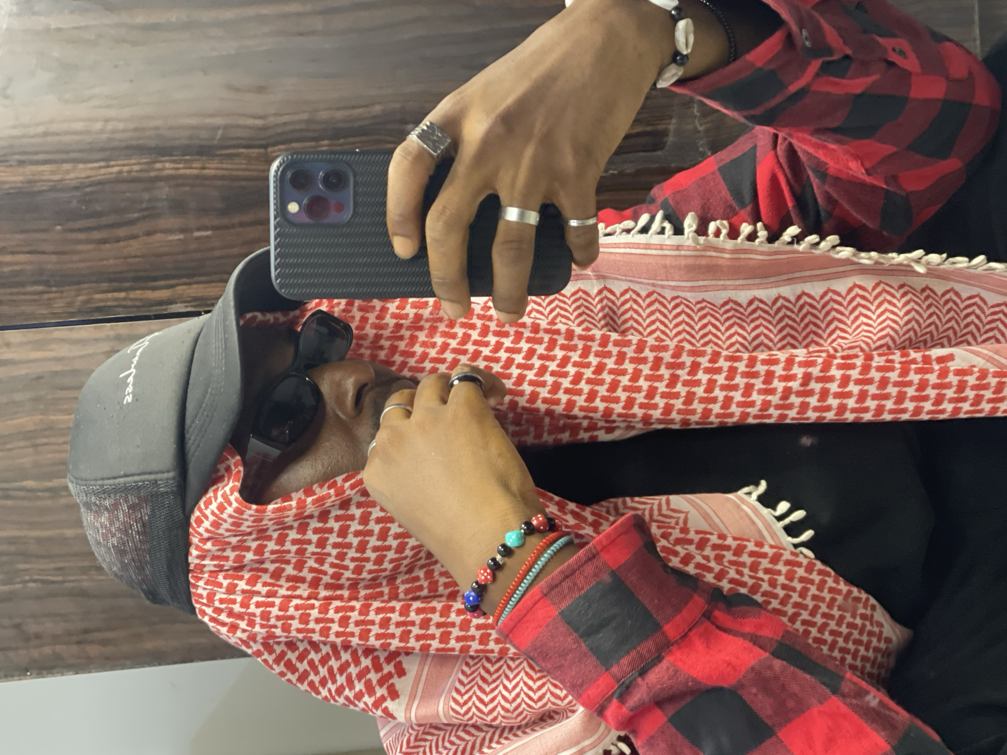 A person wearing a black mesh trucker hat, black sunglasses, a red and black plaid shirt, and a red and white keffiyeh takes a selfie in a room. The keffiyeh is draped over their head and shoulders, partially obscuring their face. They are holding a black smartphone in their right hand, which is angled to capture the reflection in a wooden-paneled wall behind them. Several silver rings are visible on their fingers, along with beaded bracelets on their left wrist. The person's left hand is resting on their chin. The overall scene suggests an indoor setting, possibly a residential or hotel room in Abuja, Nigeria, given the stylistic elements and context.