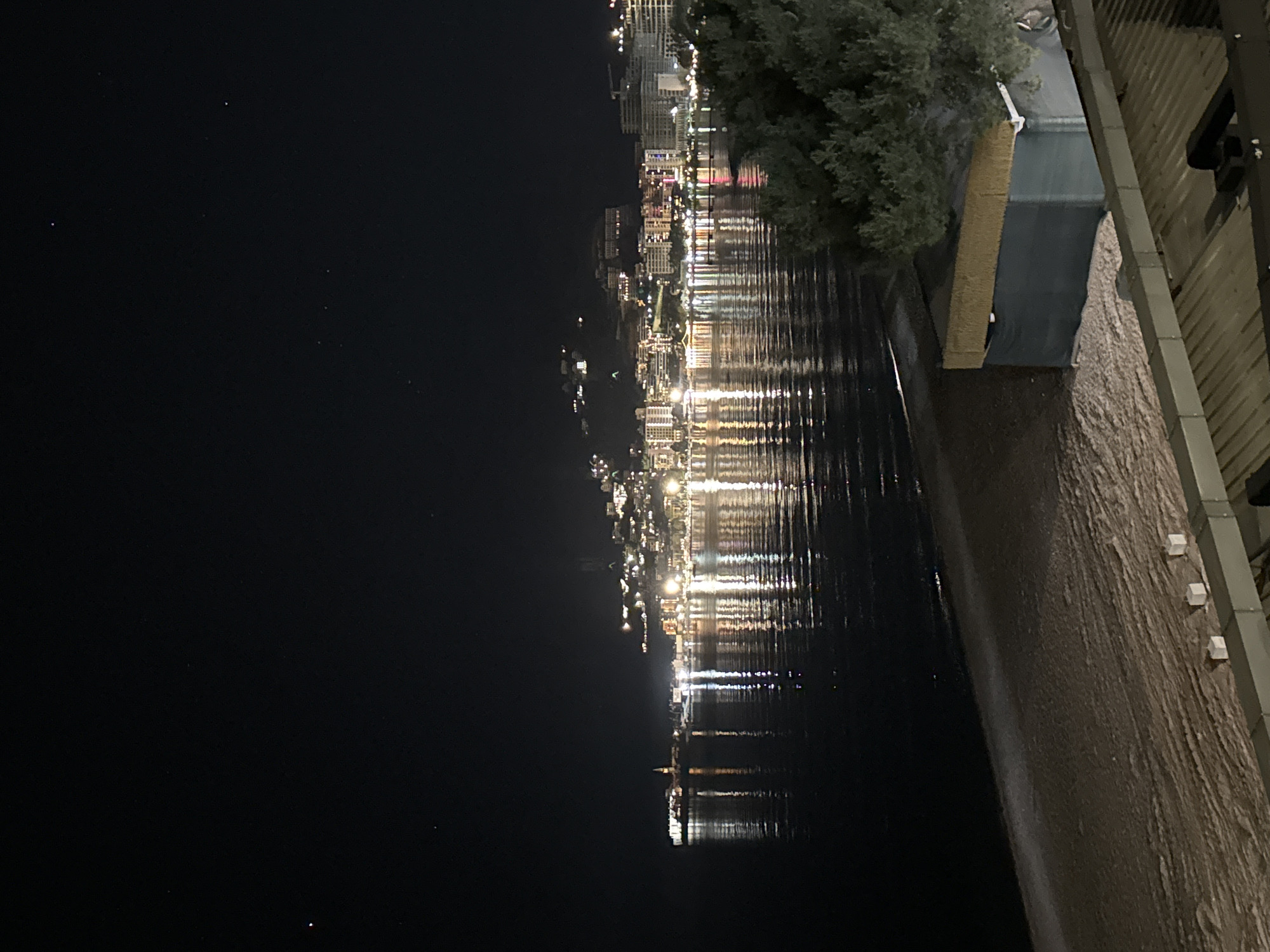 At night, the coastal city of Budva, Montenegro is captured from an elevated perspective. The left side of the frame is dominated by a dark sky, contrasting with the city lights reflected on the water. A shoreline runs along the right side, with structures like buildings and possibly trees visible at the edge. A segment of a building structure is in the right top corner of the image.
