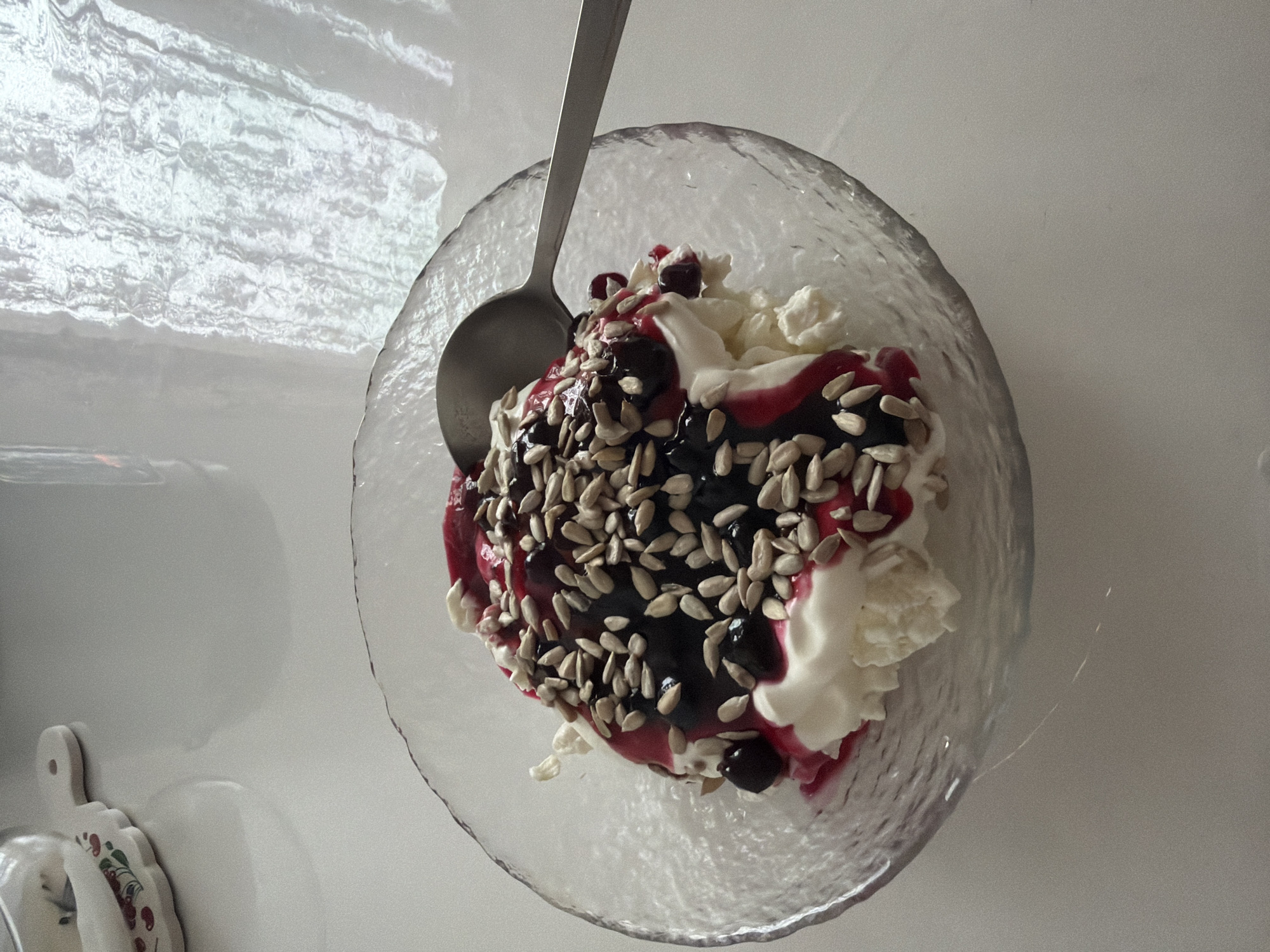 A top-down shot depicts a clear, textured glass bowl containing a dessert. The dessert consists of what appears to be a base of cottage cheese or cream, topped with a dark, glossy sauce with visible red streaks, likely berry sauce. The dessert is heavily garnished with sunflower seeds. A silver spoon rests on top of the dessert. The background is a white surface. The image was likely taken in a residential setting. There is no confirmed connection to Leningradskaya, Russia.