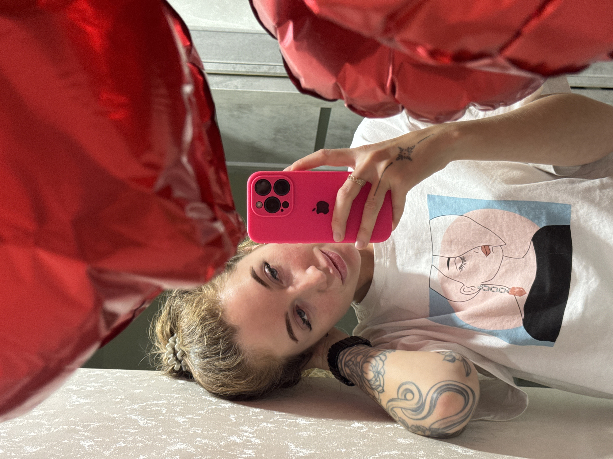 A young woman with blonde hair and blue eyes is lying down and taking a selfie with a pink smartphone. She is wearing a white t-shirt with a graphic print. Her left arm is visible and has tattoos. She is holding the phone in her right hand, which also has a tattoo and a ring. There are red balloons in the foreground. The location is Chaikovsky, Russia.