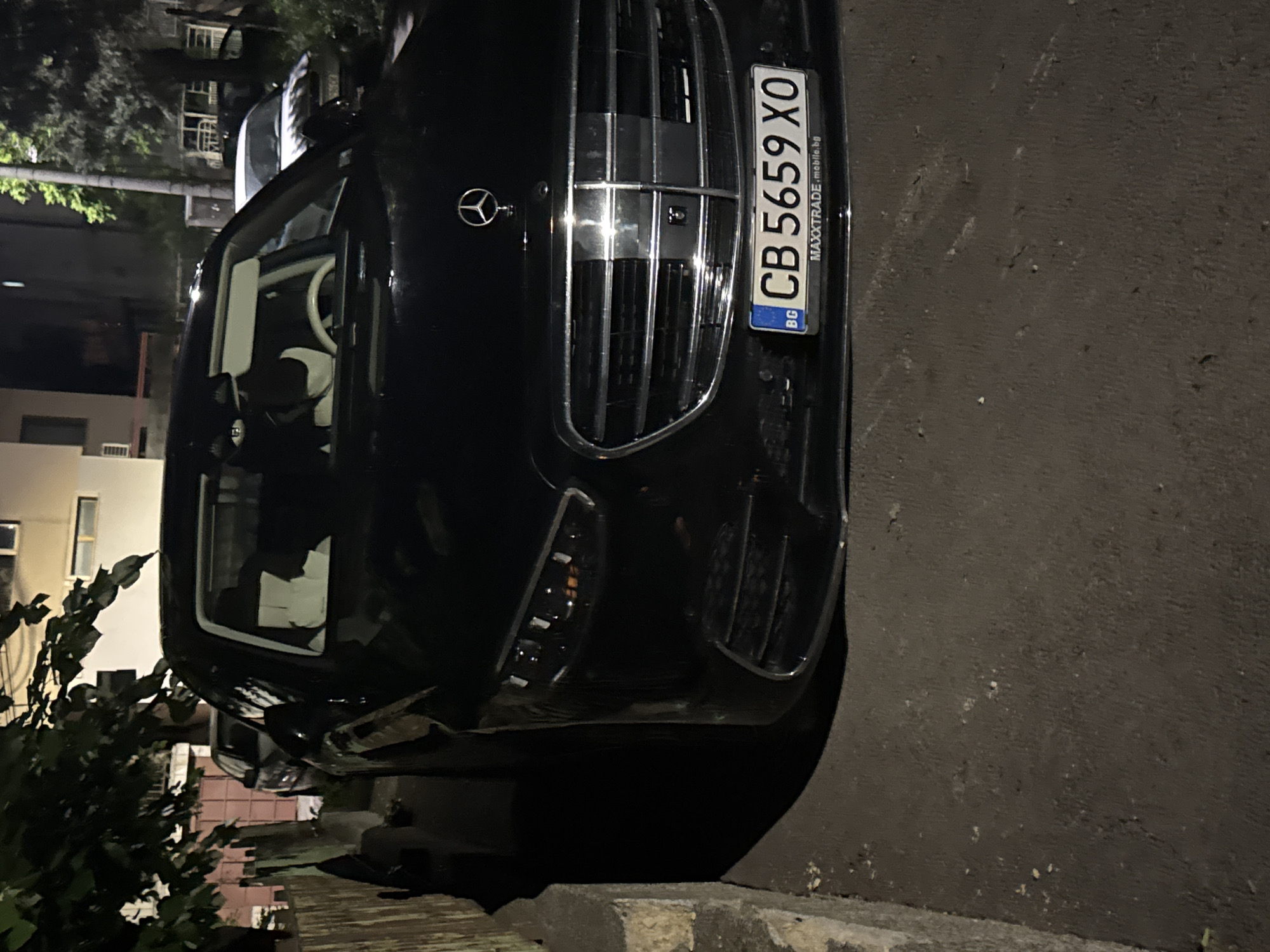 The image depicts a black Mercedes-Benz car parked on a street in what appears to be a residential area in Sofia, Bulgaria. The car is positioned close to the curb, with a portion of the vehicle visible from the front. The license plate, partially visible, reads "CB 5659 XO" and includes the "BG" country code. The background shows buildings and trees, indicating an urban setting. The lighting suggests it is nighttime.