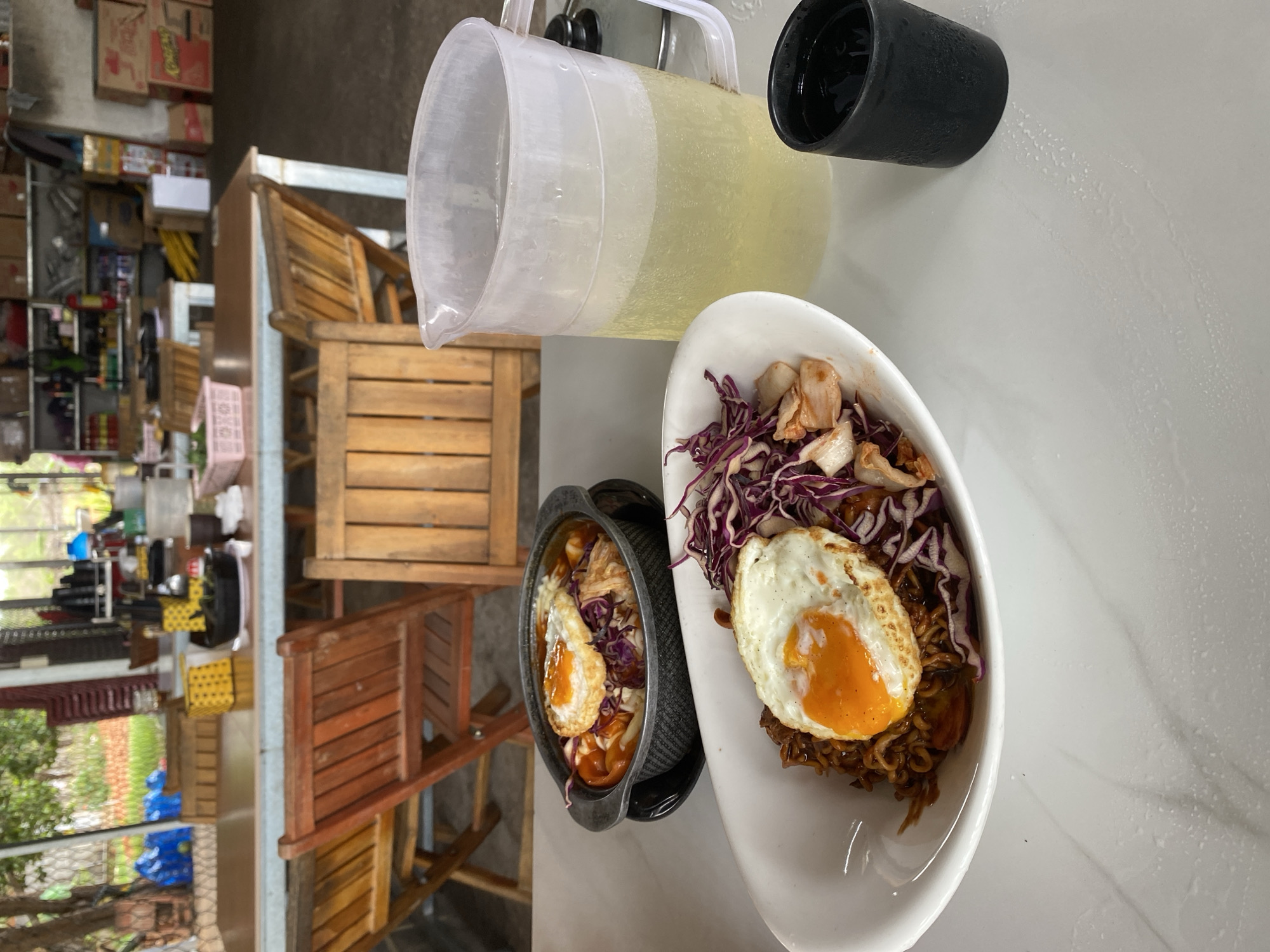 The image depicts a tabletop scene, likely within a dining area in Mũi Né, Vietnam. The tabletop is made of a white material with a marble-like pattern. A clear plastic pitcher containing a pale yellow liquid and a black drinking glass are placed on the table. There are two dishes that feature an egg with a bright orange yolk, noodles, red cabbage and other foods. A metal cooking pot with a black exterior is also present.
Wooden furniture is visible in the background. The background includes a window overlooking foliage. Boxes are stacked on a shelf in the background.