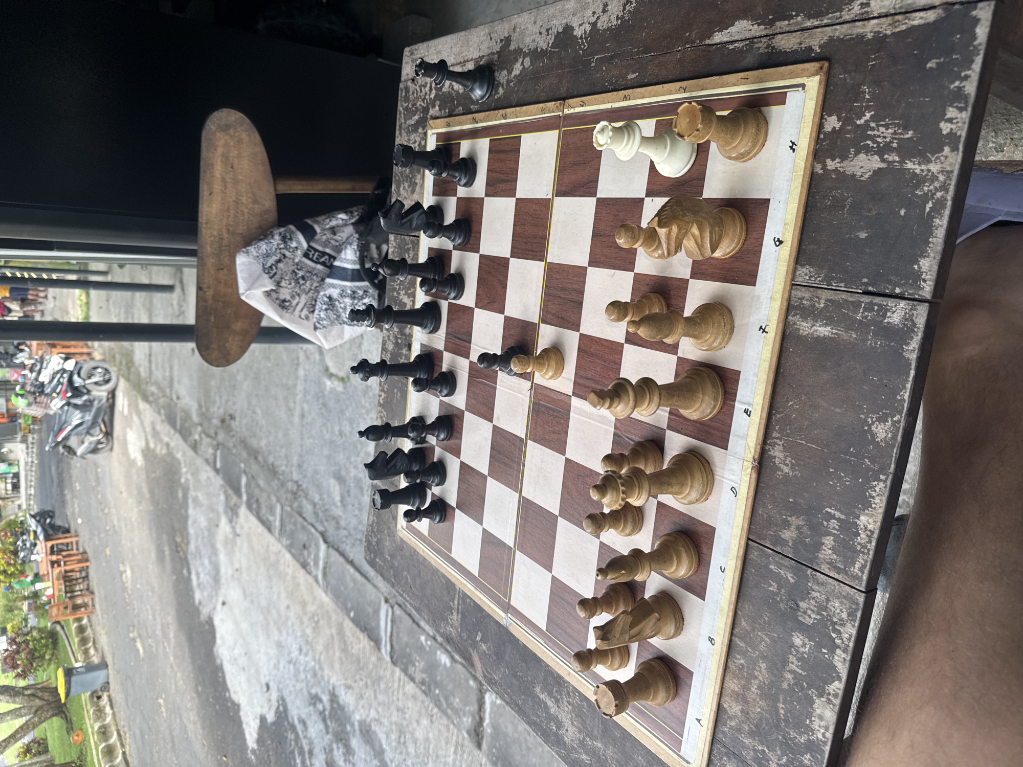 The image shows a chess game in progress on a wooden chessboard placed on a weathered wooden table. The white pieces are positioned near the bottom edge of the frame, while the black pieces are opposite them. Some pieces are slightly out of place.
Outside the partially visible window, a street scene in Kuta Selatan, Indonesia, unfolds. The street is lined with motorcycles, shops, and greenery.
A wooden stool sits adjacent to the table.