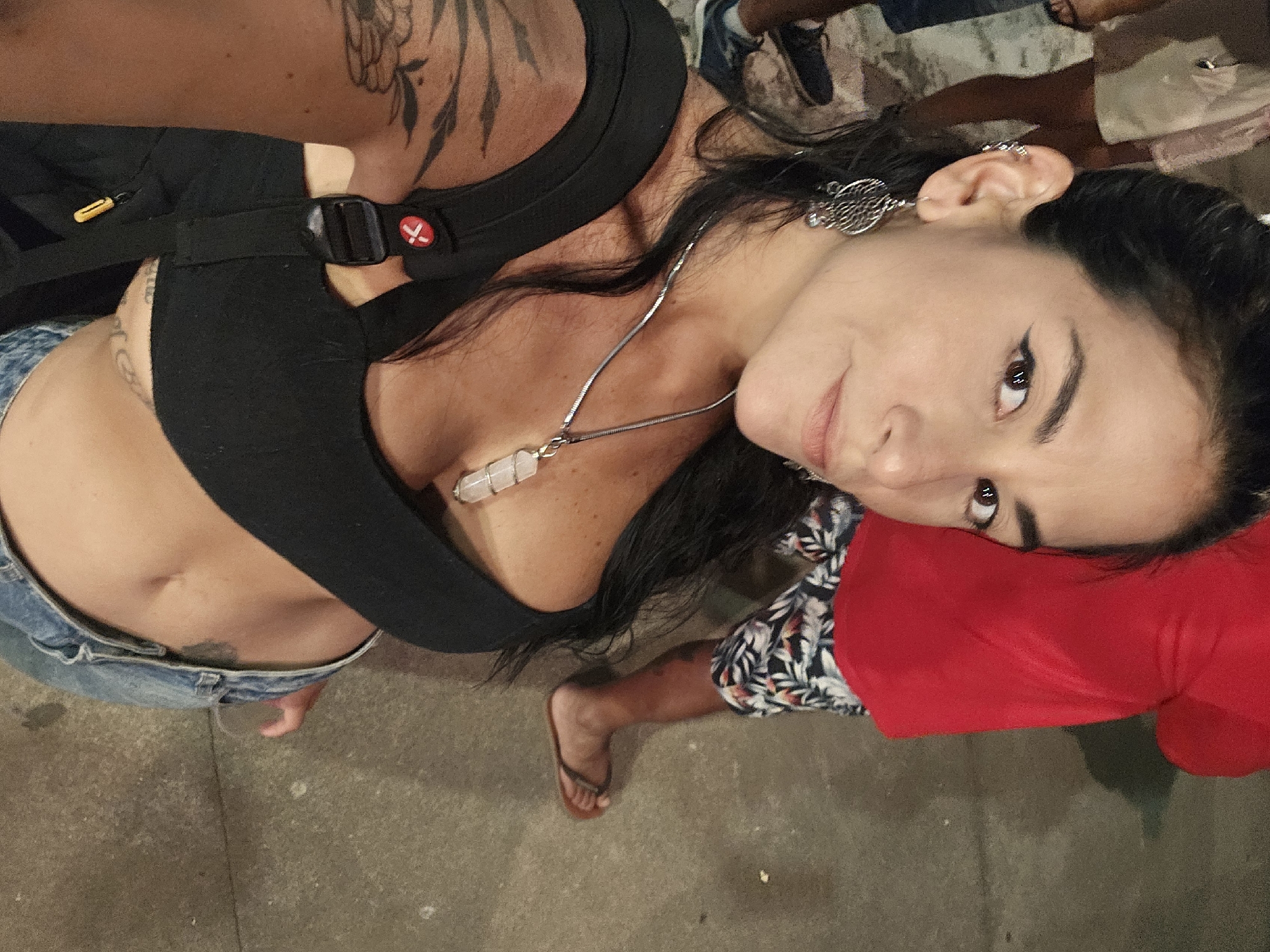 A woman with dark hair, wearing a black top, denim shorts, and a silver necklace with a crystal pendant, is the central subject of the image. She is looking up at the camera with a slight smile. Her left ear is adorned with a large silver earring, and there's a smaller earring further up her ear. Parts of tattoos are visible on her chest, stomach, and shoulder. She is wearing a black harness over her top. The ground is a grayish concrete. Parts of other people are visible, including a foot in a flip-flop, and clothing in floral and red patterns. Location: Armação dos Búzios, Brazil cannot be determined from the image.