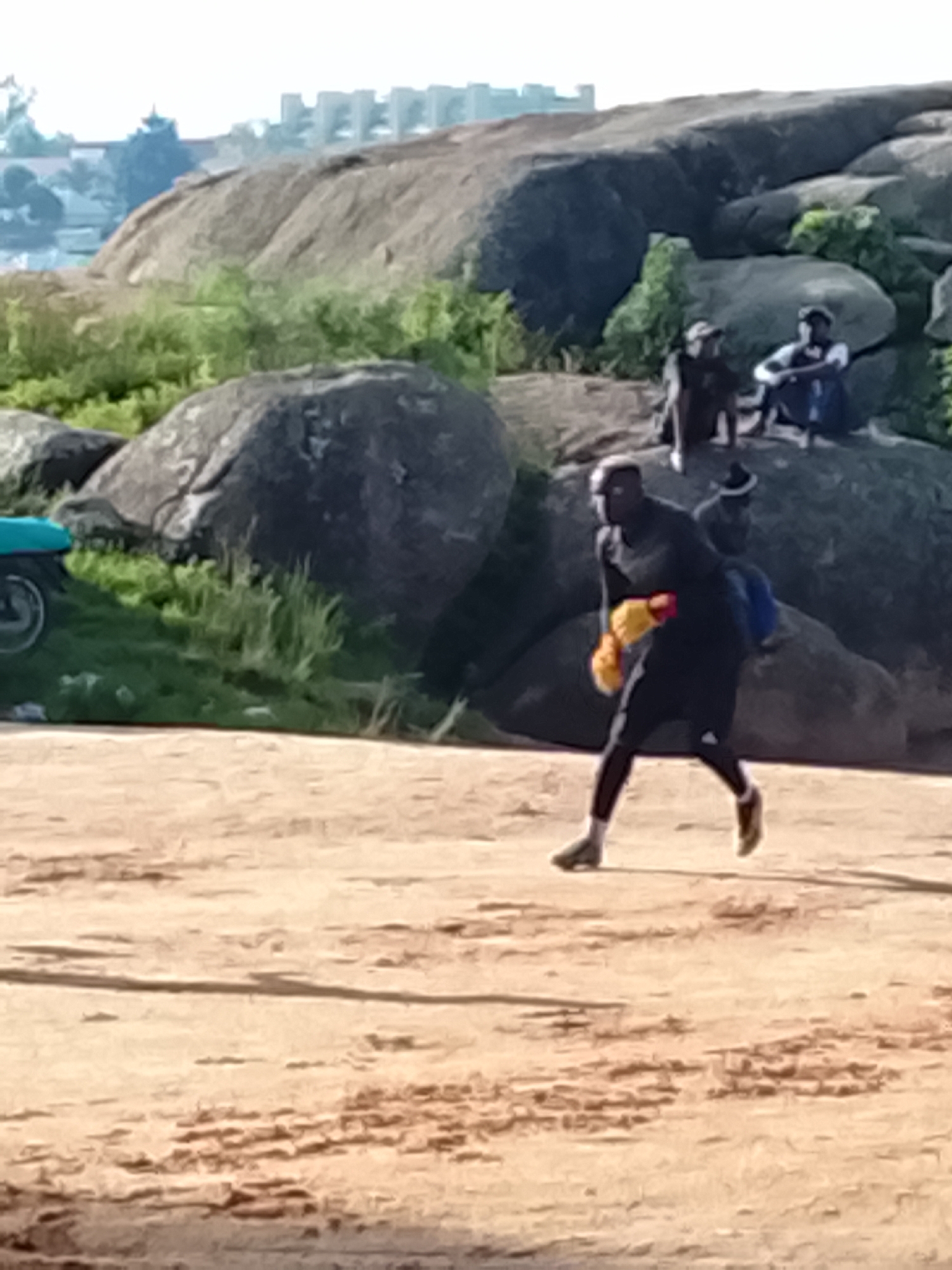 In Jos, Nigeria, the image depicts a group of individuals in an outdoor setting characterized by large rock formations and a sandy ground. A man is prominently featured in the foreground, actively engaged in boxing training. He wears a black outfit, including a headguard, and yellow boxing gloves. His posture suggests movement, possibly a punch or a defensive stance. In the background, several other individuals are seated or standing on the rocks. One person appears to be wearing a blue shirt. The environment is dry and appears to be arid, with sparse green vegetation growing amongst the rocks. A portion of a blue vehicle is visible on the left side of the frame, partially obscured by green foliage. In the distance, buildings can be seen, suggesting the proximity of an urban area. The lighting indicates it is daytime, with the sun casting shadows.