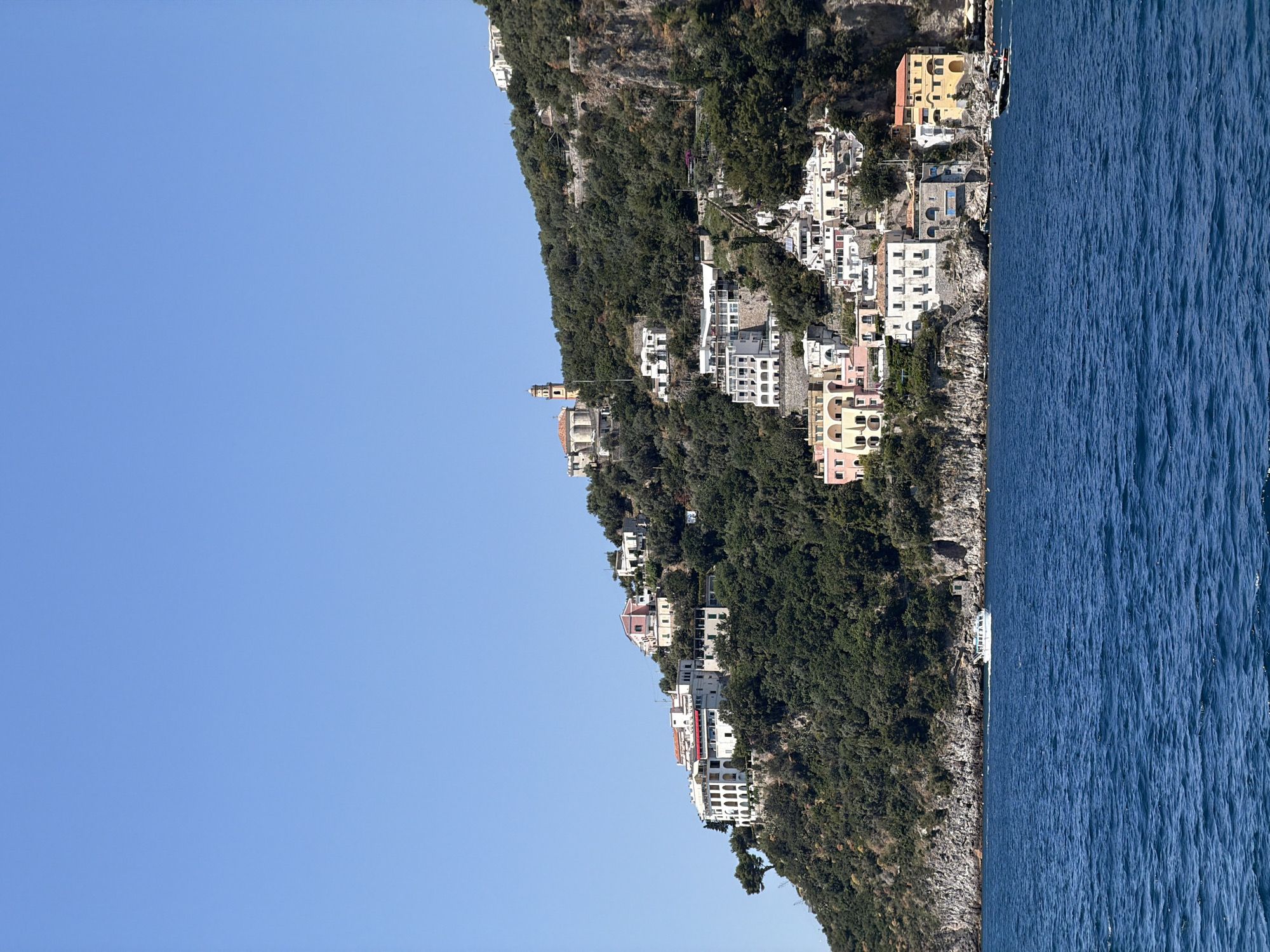 The image depicts a coastal Italian landscape with a clear blue sky on the left and the rippling surface of the sea on the right. A steep cliff dominates the center, densely covered with green vegetation, and dotted with colorful buildings reminiscent of the Amalfi Coast, Italy. The buildings vary in size and style, clinging to the hillside and suggesting a tiered urban layout. A church steeple is visible higher up on the cliff.