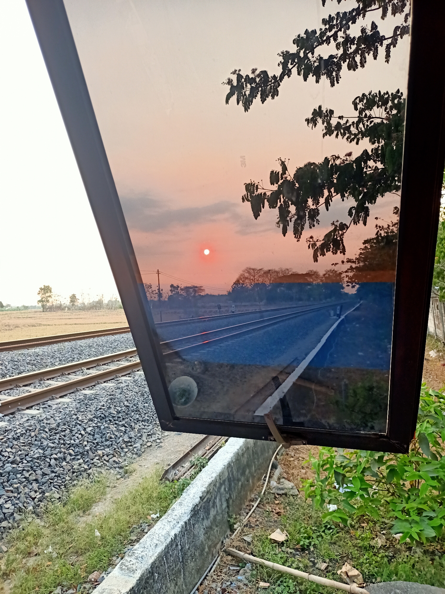 This image captures a view of railway tracks in Banjarjo, Indonesia, as seen through a reflective surface, likely a window or a sign. The foreground shows gravel ballast supporting two sets of railway tracks. To the left, a field of dry grass extends towards a line of trees in the distance. A concrete structure with white paint runs along the edge of the tracks.
The reflective surface, positioned at an angle to the viewer, displays a sunset scene. A small, bright sun hangs in a pale orange sky, partially obscured by faint clouds. Silhouetted branches and leaves of a tree are visible against the sky, with some appearing to be in the foreground of the reflection, suggesting they are closer to the reflective surface. The reflection also shows the railway tracks stretching into the distance, with a white line painted along the edge of one track. In the lower part of the reflection, a circular object, possibly a reflection of a light or a circular object mounted on the reflective surface, is visible. The overall lighting suggests it is either early morning or late afternoon.