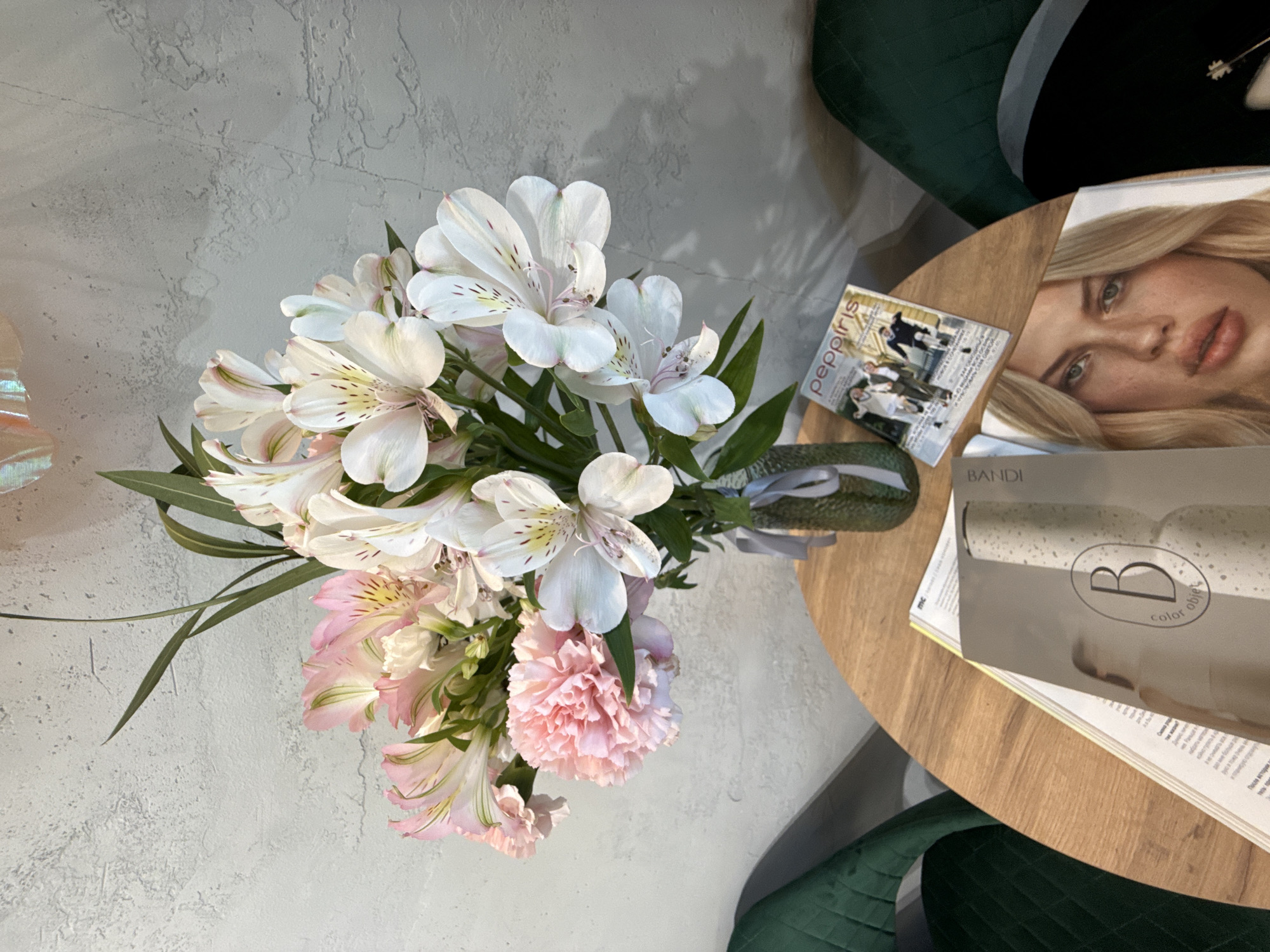 The image captures a top-down view of a bouquet of flowers. The bouquet consists of white and light pink Alstroemeria flowers, also known as Peruvian lilies, and a pink carnation. The flowers are arranged in a vase with a green textured exterior and tied with a gray ribbon.
The bouquet sits on a light-colored, textured surface, likely a concrete or plaster wall. To the right of the bouquet, the edge of a round, light-wood table is visible. Stacked on the table are magazines or booklets. One magazine features a blonde woman on the cover. Another magazine is visible with the brand name "BANDI" and a logo including the letter "B" within a circle. A copy of "peppiris" magazine can be seen as well.
The corner of two dark green chairs is visible to the right and bottom right corner.
Given the presence of the Russian-language magazine "peppiris," it is plausible that this image was taken in Russia, but without further clues, specifying Saint Petersburg is not verifiable from the image alone.