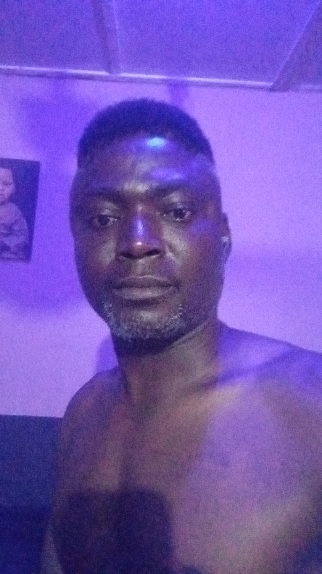 This media file is a portrait of a man, likely taken indoors under artificial lighting. The subject is an adult male with dark skin, short dark hair, and graying stubble on his chin and jawline. He is shirtless, with his shoulders and upper chest visible. He looks directly at the camera with a neutral to slightly serious expression.
The entire scene is bathed in a distinct purple or violet light, which gives the image a monochromatic feel. This lighting reflects off the man's skin, making parts of his face appear somewhat shiny. In the background, to the left, a framed picture of what appears to be a child's face is visible on a wall. Above, a ceiling or structural element is present. The image has a slightly grainy texture. There is no visible text in the media file.