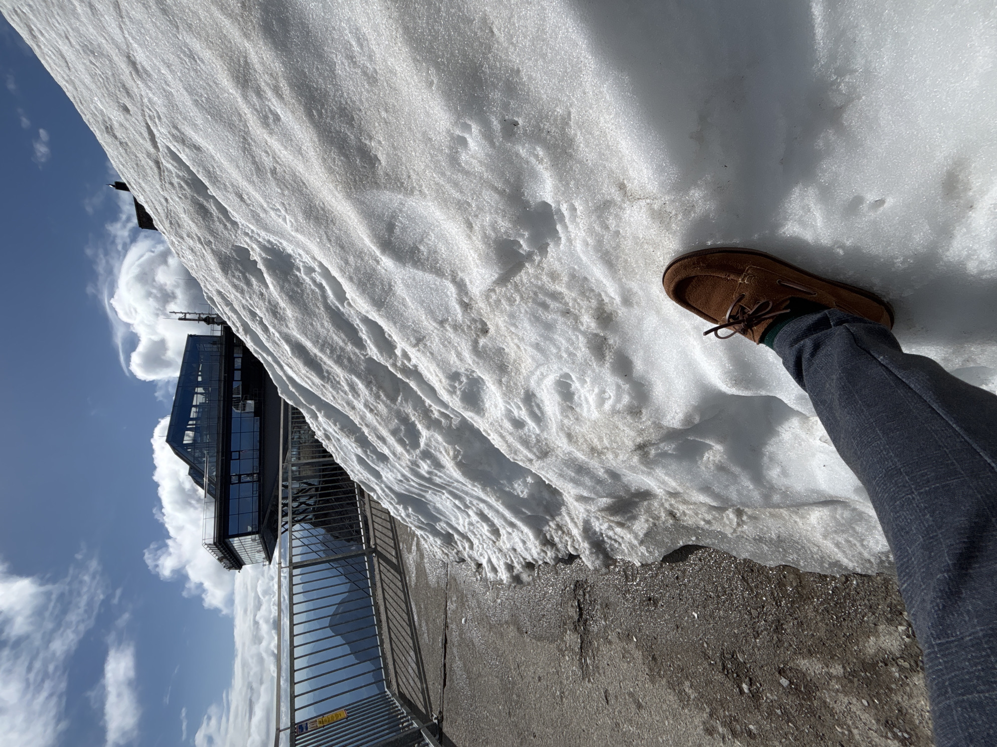 This is a first-person perspective, eye-level medium shot of a person's legs and feet near a large snowdrift at the Ehrwald, Austria. The person is wearing dark gray checkered trousers and a brown suede boat shoe with green socks. The snowdrift occupies the right two-thirds of the frame and shows signs of melting, with some patchy dirt and water visible at its base. To the left of the snowdrift, a paved walkway is visible, speckled with dirt and gravel, and wet in places. A metal railing runs along the edge of the walkway, separating it from a steep drop-off. In the background, a modern building with large glass windows, a restaurant or observation deck, is perched on the mountainside. The sky above is blue with scattered white clouds. The scene appears to be a scenic mountain location.