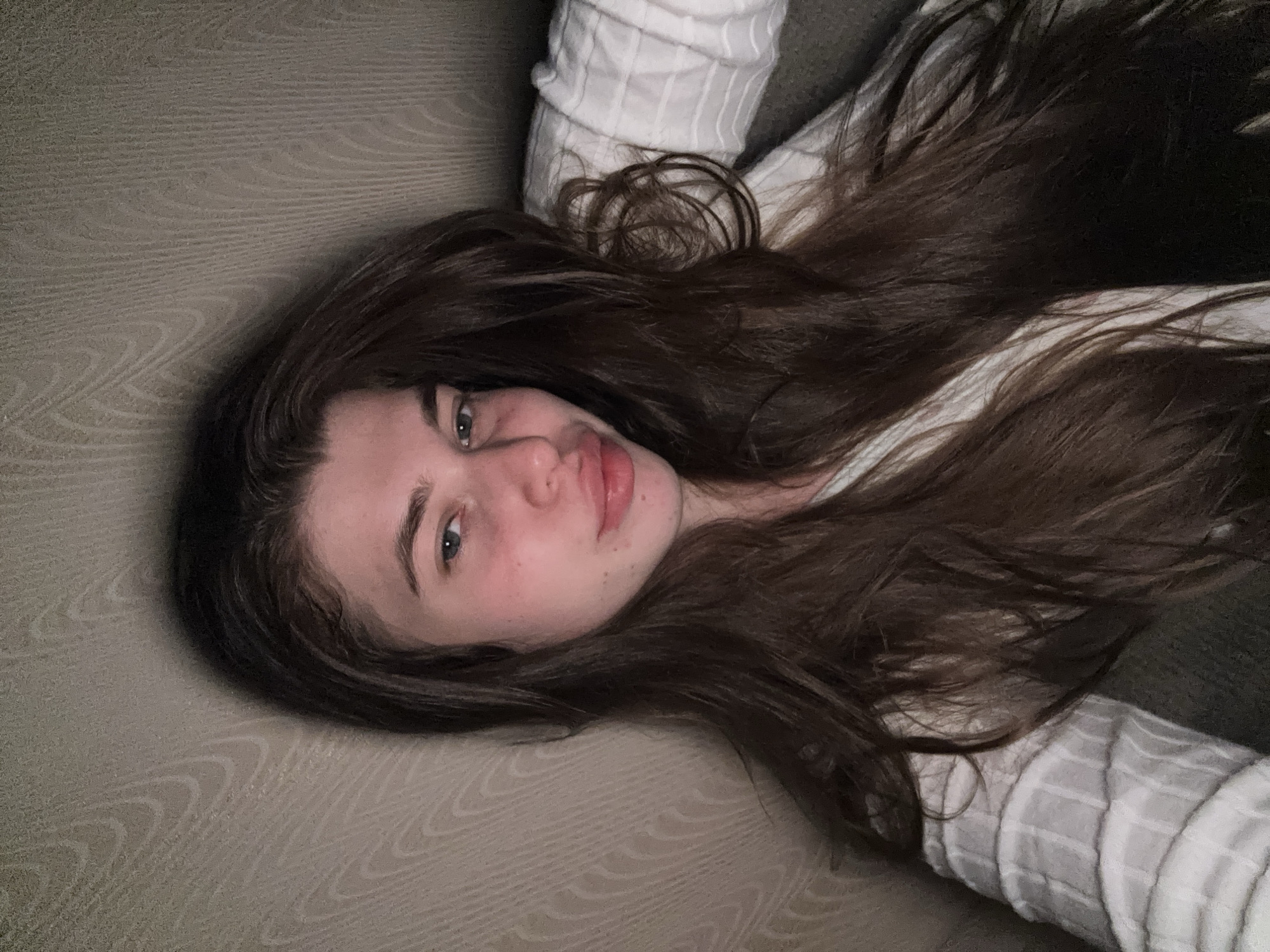 A young woman lies on a gray surface, likely a couch or bed, with her dark brown hair spread around her head. She is wearing a white shirt with a subtle striped pattern. Her face is visible, with fair skin, light blue eyes, and slightly parted lips. The lighting is dim and diffused. The location is not discernible as Мещёры, Russia, or any specific city. The focus of the image is the woman's face and hair.