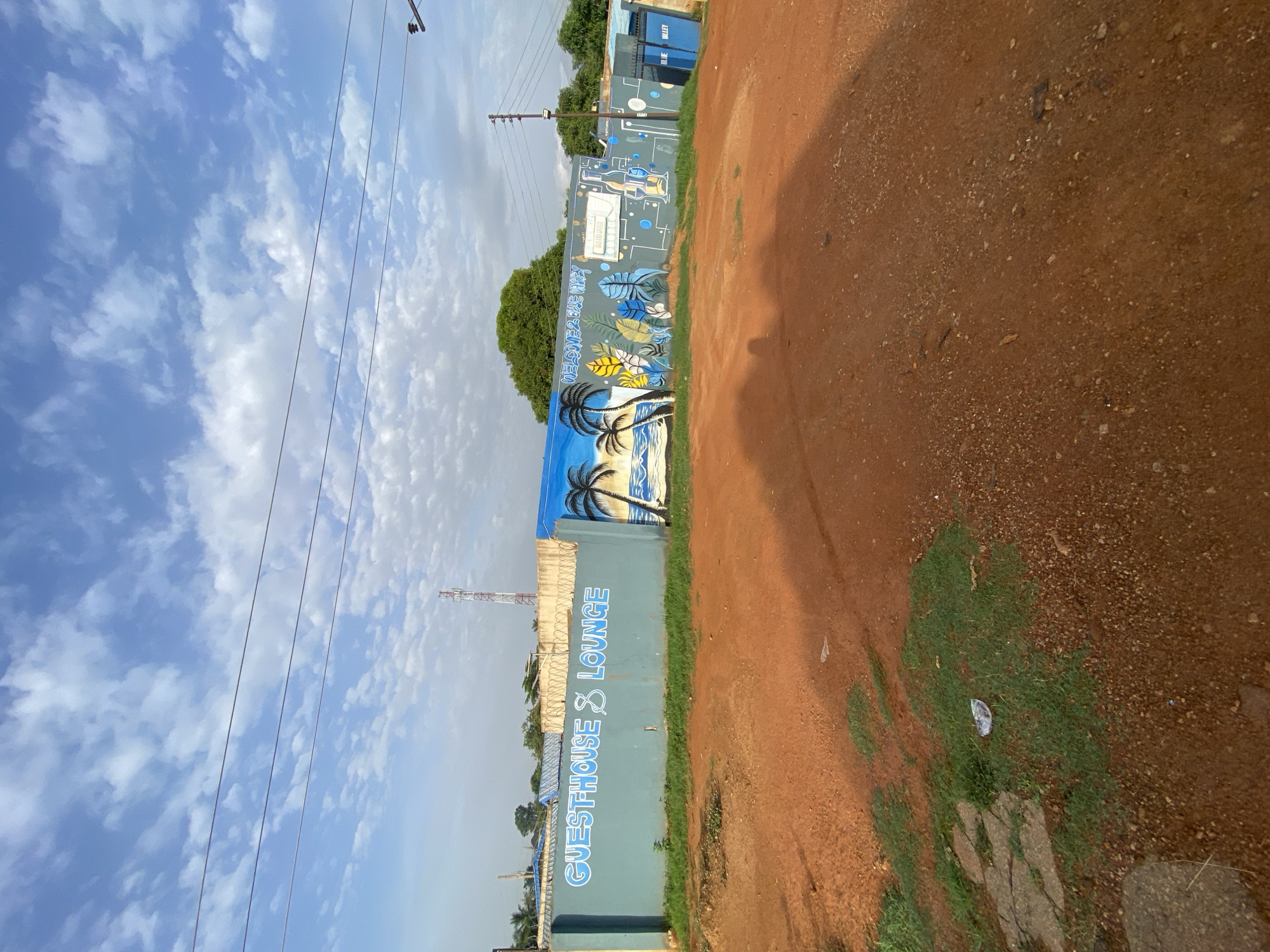This is a wide, eye-level shot of a blue-painted concrete wall with a mural on it, located in Bukuru, Nigeria. The wall is approximately 10 feet high and stretches across the frame. The mural depicts a beach scene with palm trees, waves, and a blue sky with white clouds. The words "GUESTHOUSE & LOUNGE" are written in large, white, sans-serif font on the left side of the wall. To the right of the beach scene, there is text that reads "WELCOME TO BLUE VALLEY" and further to the right are drawings of tropical leaves, bottles, and glasses. A blue metal gate with the words "BLUE VALLEY" on it is visible on the far right of the wall.
In the foreground, a dirt road made of reddish-brown soil occupies the lower half of the image. Patches of green grass and a few pieces of white litter are scattered on the dirt. In the background, above the wall, a communication tower with red and white sections is visible, along with some trees and a bright blue sky with scattered white clouds. The lighting suggests it is daytime, with shadows cast on the dirt road from an unseen object to the right.