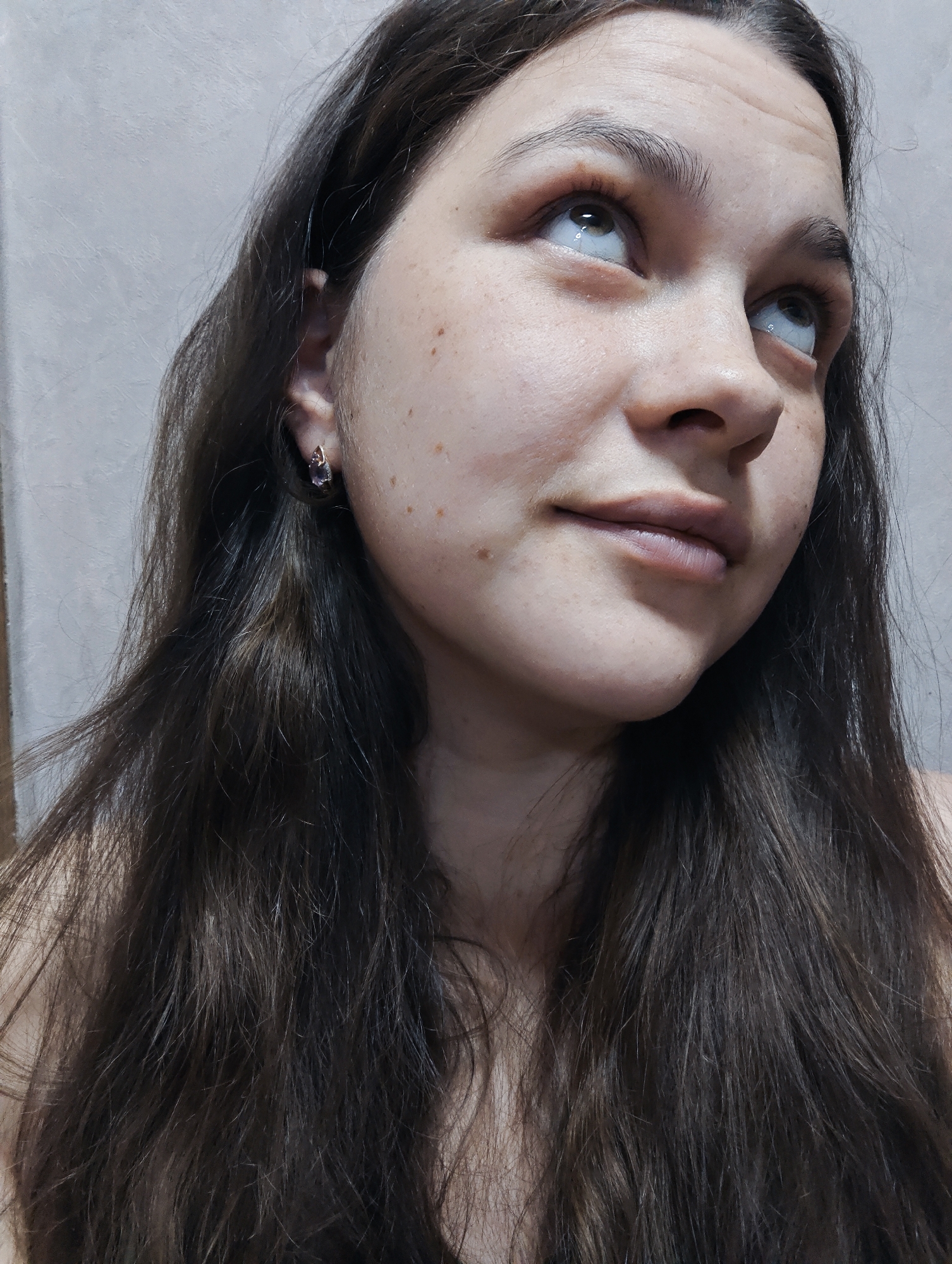 The image features a young woman from the chest up, looking upward and slightly to her right. She has long, dark brown hair, parted on the left, which falls over her shoulders and obscures most of her left side. Her skin appears fair with numerous freckles visible across her cheeks and forehead. She has a subtle, almost Mona Lisa-like smile on her lips. Her eyes are light in color, possibly hazel or green, and appear to be wet or reflective as she gazes upwards. A small, decorative earring with a purplish stone is visible in her right ear. The background is a plain, light grey or beige wall with a slightly textured finish, suggesting an indoor setting. There is no visible text in the image. The overall impression is one of thoughtfulness or gentle amusement.
