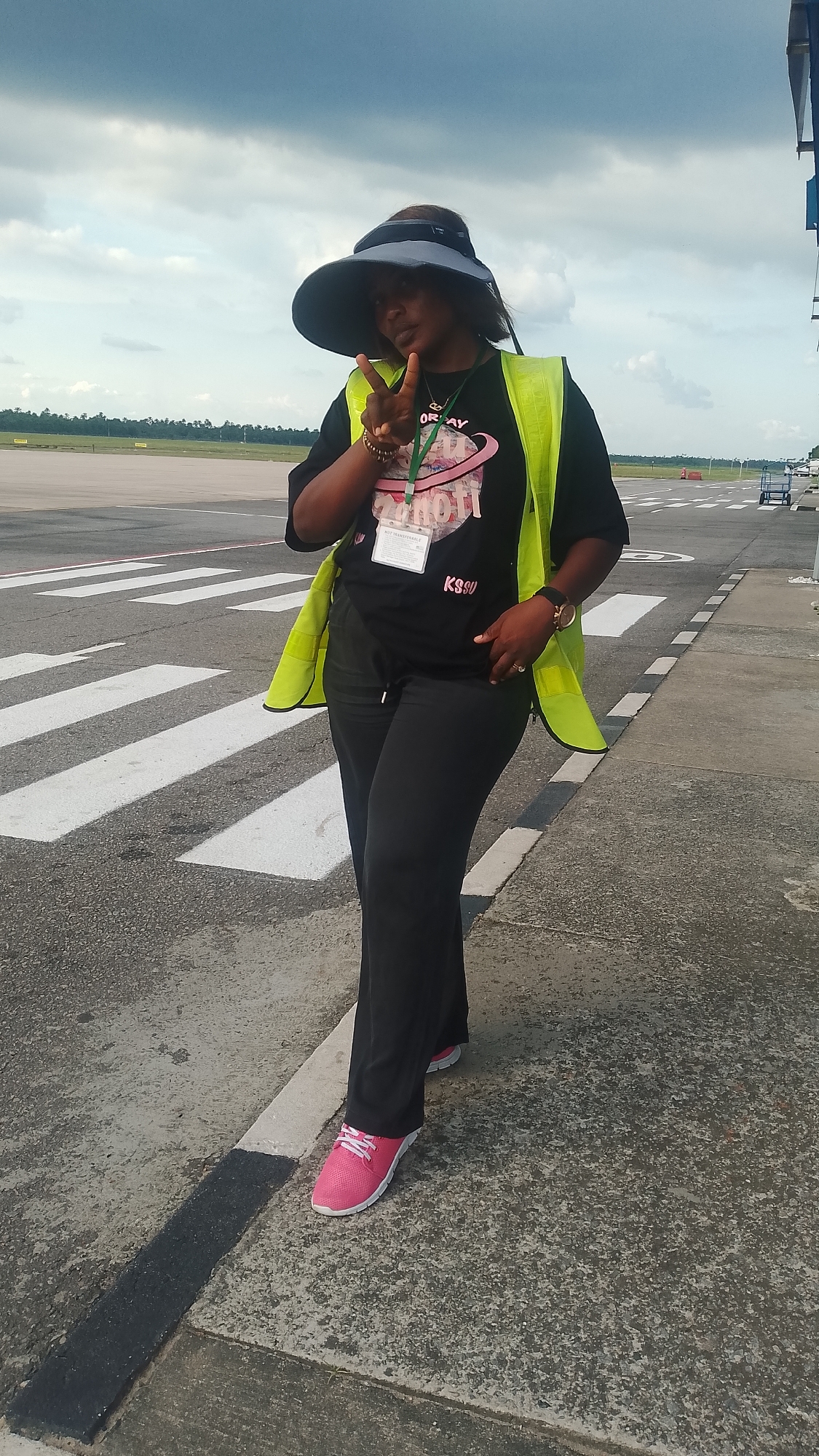 A woman stands on the edge of an airport runway in Orerokpe, Nigeria, near a crosswalk, giving a peace sign with her right hand. She wears a black t-shirt with a graphic design, black pants, pink sneakers, and a large sun visor. A yellow safety vest is worn over the shirt. She has a lanyard with a name tag around her neck. The runway stretches into the distance under a cloudy sky. The ground is paved and marked with lines. A luggage cart sits in the distance.