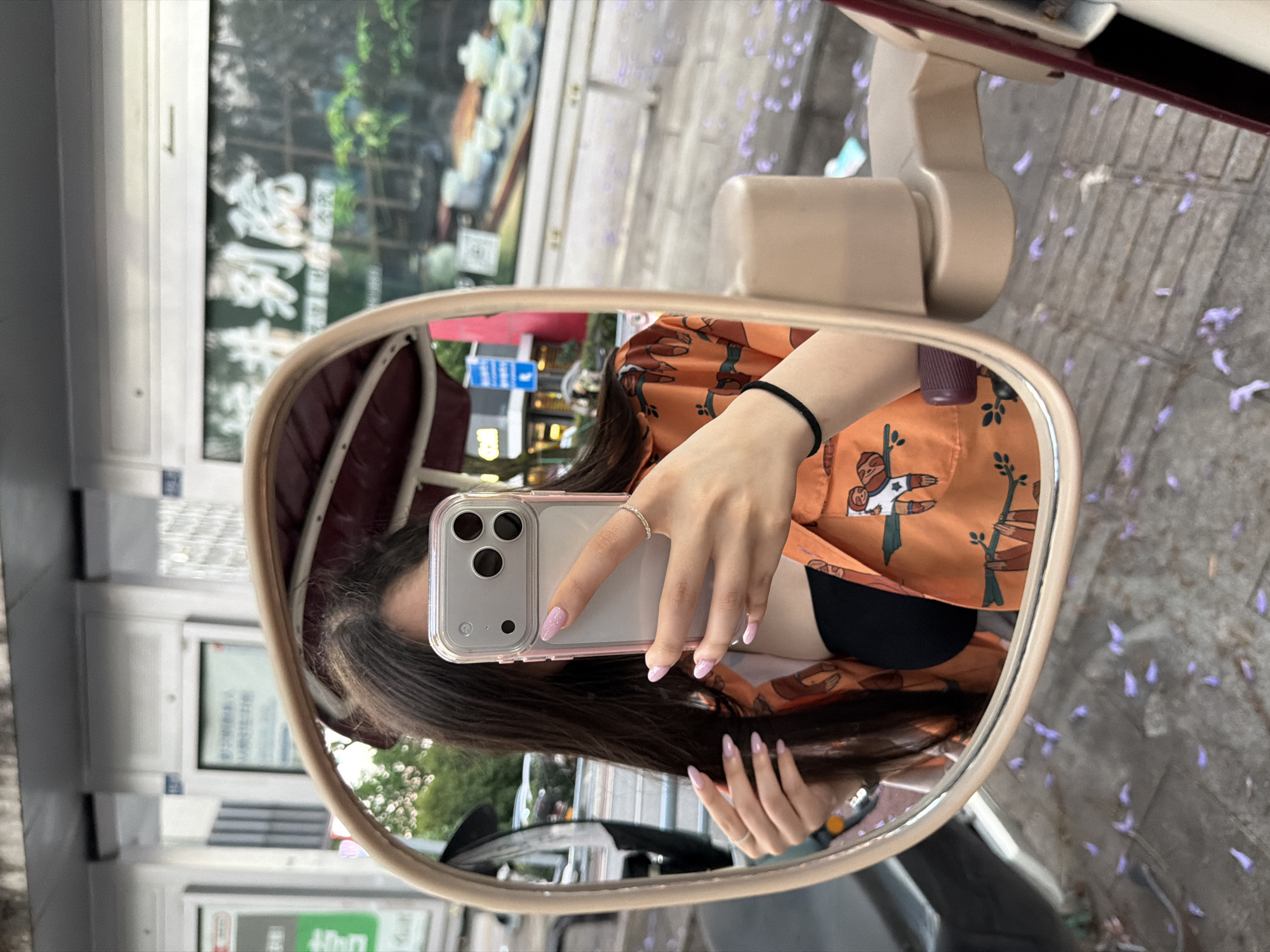 A mirror's reflection shows a person holding a smartphone, with long dark hair, wearing an orange patterned shirt and a black top. The mirror is attached to a beige vehicle accessory. The background reflects an urban environment. In the foreground are purple flowers scattered on a paved surface. The photo was taken in the city of Chongqing High-tech Zone, China.
