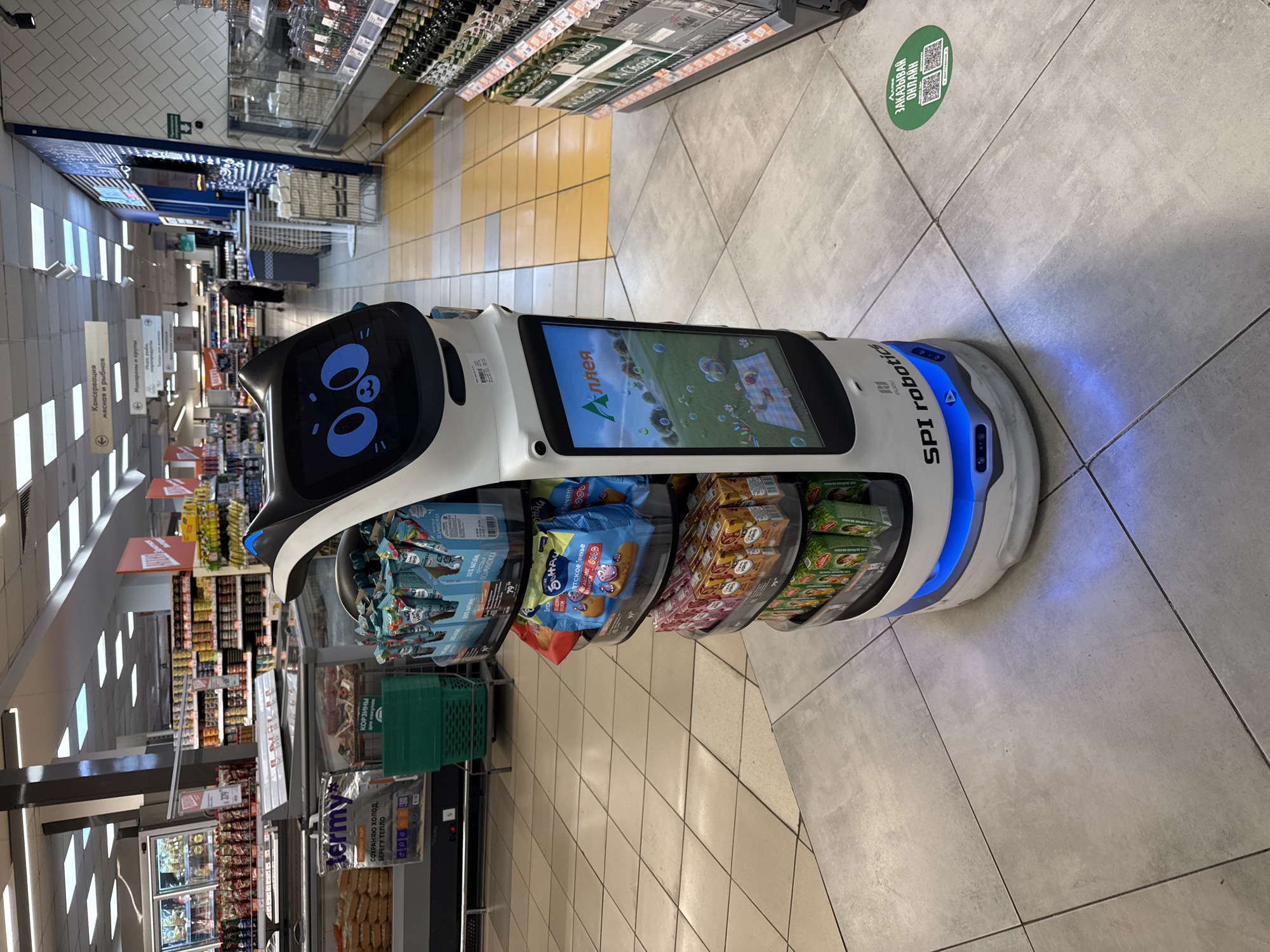 The image shows a promotional retail robot in a supermarket aisle in Krasnoyarsk, Russia. The robot, branded by "SPI robotics" and featuring a cartoon cat face and a screen displaying the "Alley" logo, is stocked with snacks on rotating shelves. Adjacent is a green shopping basket and a refrigerated display case with the "Termy" brand. Shelf signage in Russian indicates "Meat and Fish Preserves" and "Pasta and Cereals." A floor decal encourages online orders. The supermarket aisle is well-lit with tiled flooring.