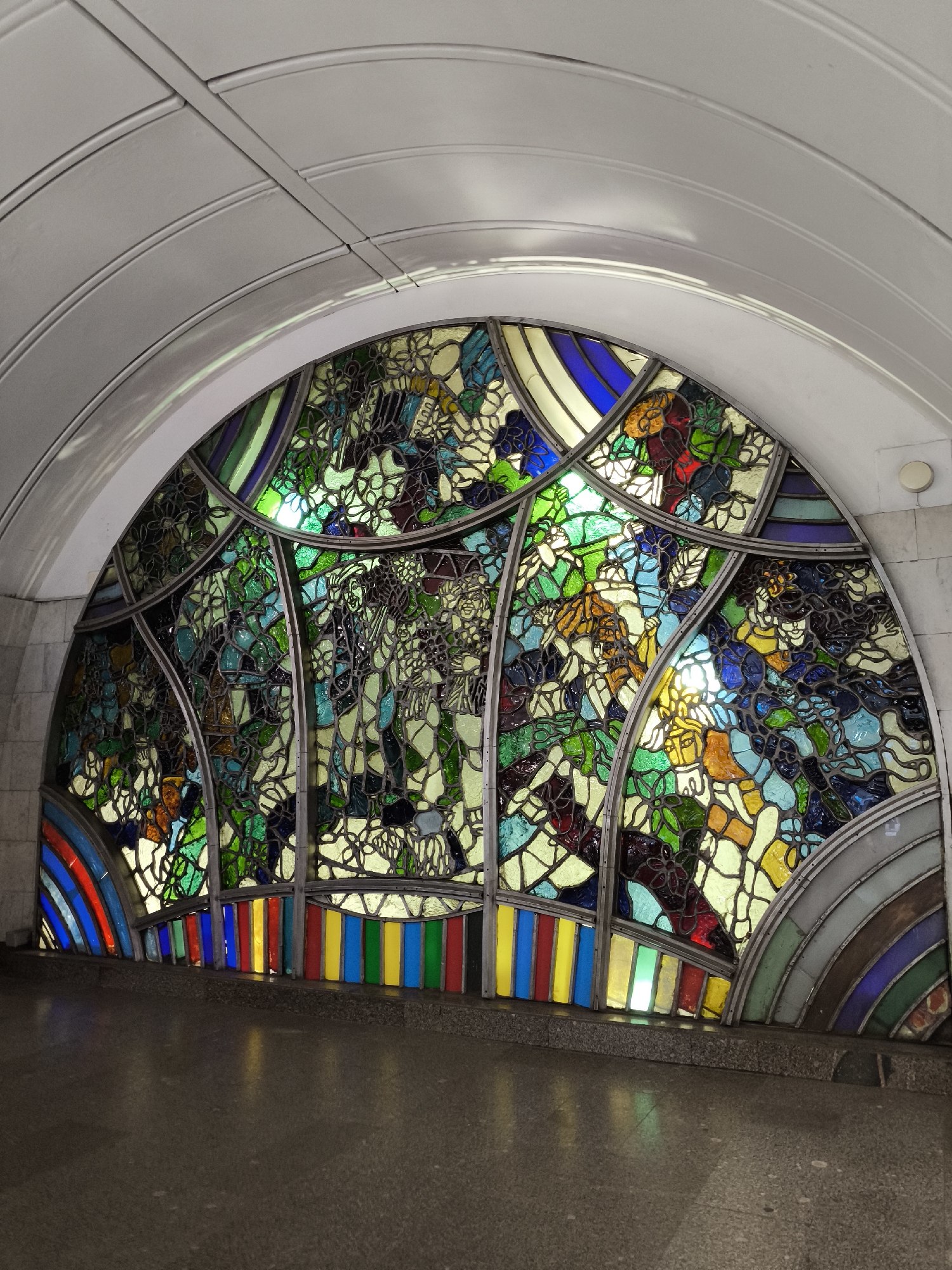 A semicircular stained glass window is set into a white, arched ceiling of what appears to be a subway station. The stained glass is vibrant and depicts figures in a stylized, artistic manner, with a dominant palette of greens, blues, yellows, reds, and white. The window is framed by metal strips. Below the stained glass, a horizontal band of colorful rectangular glass pieces runs across the bottom, resembling a rainbow or a set of colored bars, with individual pieces in red, yellow, blue, green, and white. The floor in front of the window is a polished, speckled gray material, reflecting the colors of the stained glass. The walls on either side of the window are made of light-colored, textured material, possibly marble or stone. The overall impression is one of art and public decoration within an architectural setting.