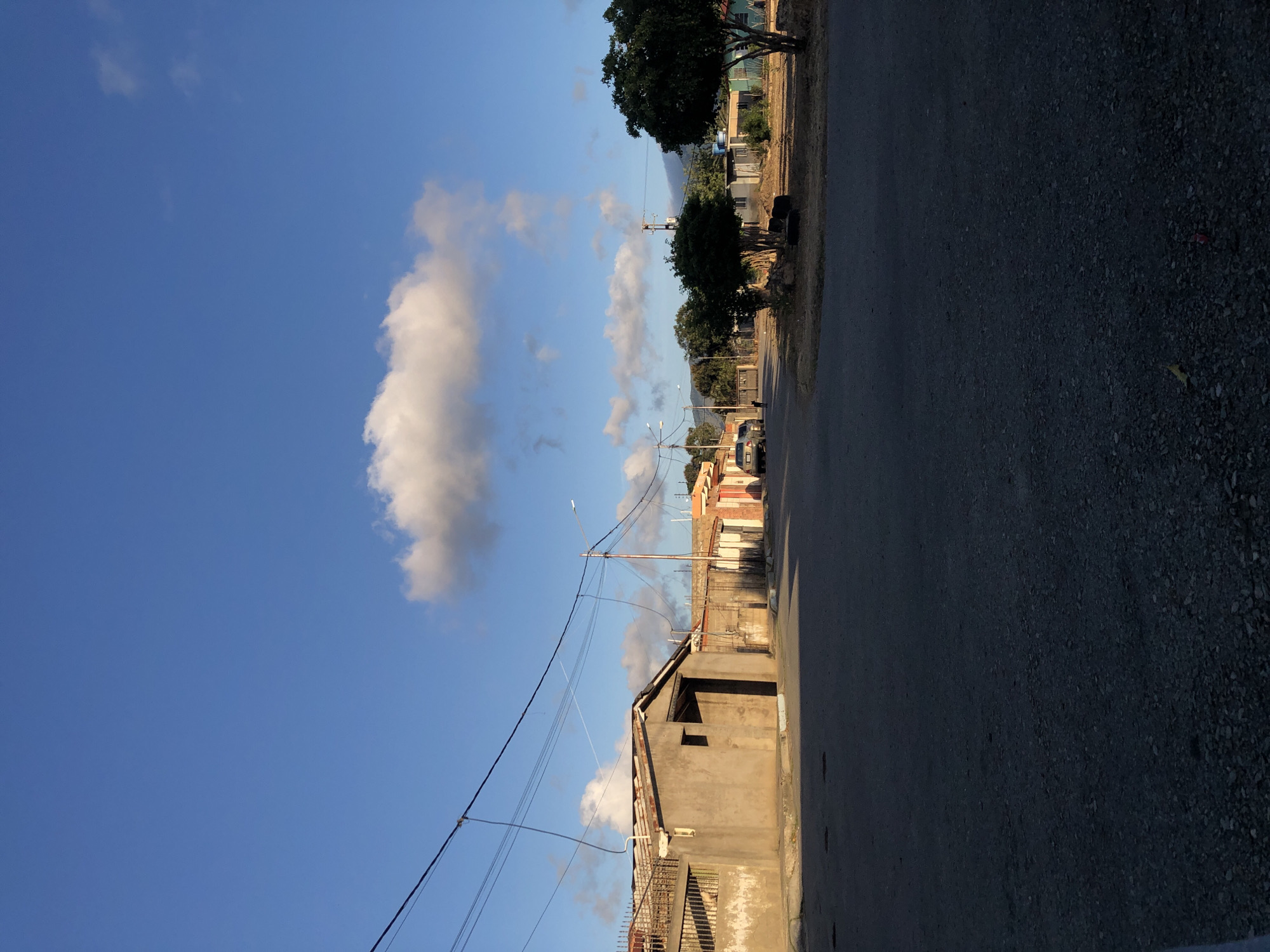 The image depicts a street scene in Altagracia, Venezuela. The sky is blue with scattered clouds. Along the right side of the frame is a road or street, with a gravelly shoulder. Buildings line the right side, varying in color from beige to reddish-orange. Utility poles and power lines run alongside the buildings. In the distance, trees are visible. No people are visible in the scene.
