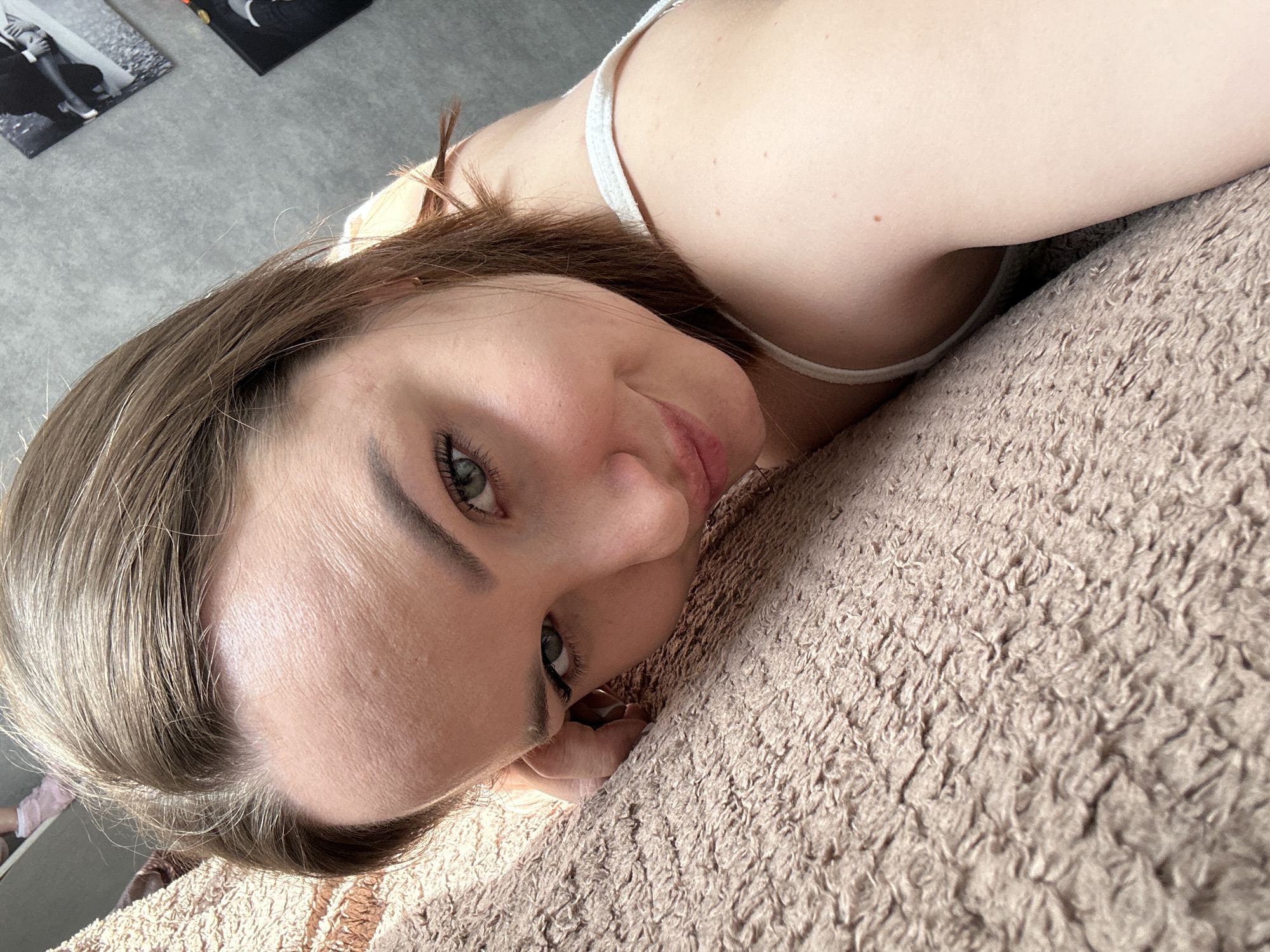 A young adult female with light skin, brown hair, and green eyes is depicted in a close-up, selfie-style photograph. She is positioned on her left side, her head resting on a light brown, shag-textured fabric surface, likely a blanket or bedspread. Her left hand's fingers are partially visible near her face, also resting on the fabric. She wears a white strap over her right shoulder, indicative of a tank top or camisole. Her gaze is directed at the camera, and she displays a slight smile. In the background, a light grey wall is visible, on which two vertically oriented black and white framed images are mounted. The scene is situated in the city of Chaikovsky, Russia.