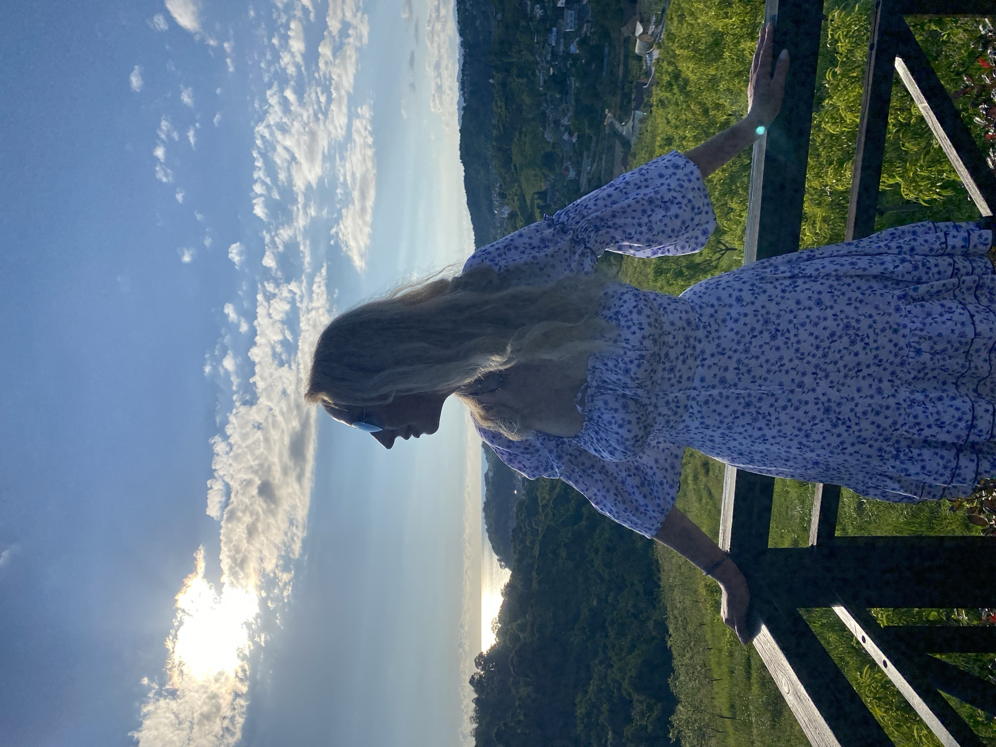 A blonde woman wearing a white dress with a blue floral pattern stands on a wooden balcony, leaning on the railing and looking out at a coastal landscape. The sun is setting, casting a warm glow on the scene and creating a reflection on the water. The background shows a lush green hillside with houses and trees, characteristic of the Sochi region in Russia. The sky is blue with scattered clouds.