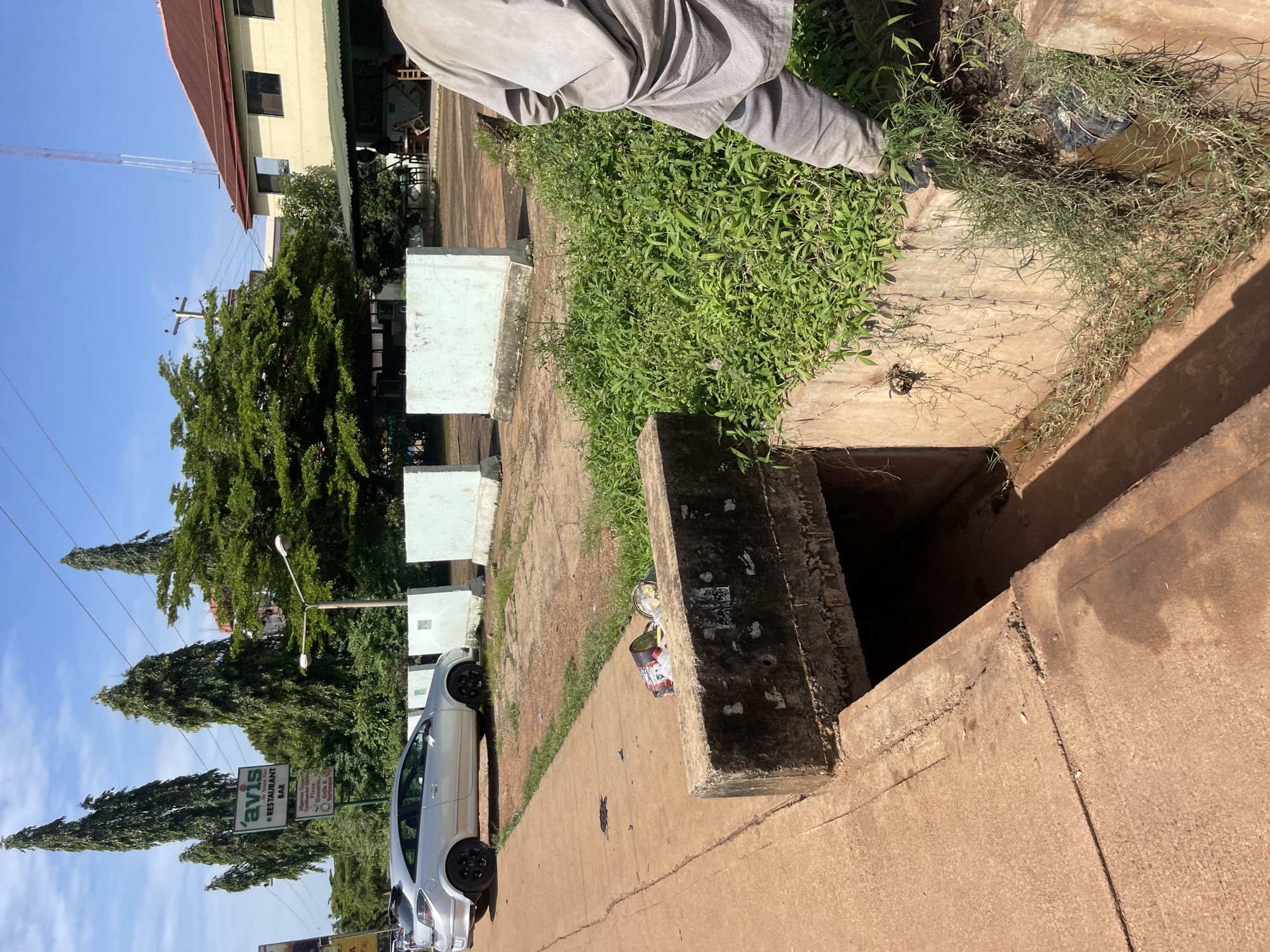 The image depicts a scene likely within the city of Jos, Nigeria, characterized by its reddish-brown soil and a mixture of urban elements and overgrown vegetation. In the foreground, a concrete drain runs along a sidewalk, with some debris and a plastic bag visible near its edge. A person, whose lower body is visible, is standing near the drain, wearing loose-fitting grey traditional attire.
In the background, a silver hatchback car is parked on the street. A prominent sign for "Avis Restaurant Bar" stands tall, surrounded by tall, slender trees. Further in the background, a multi-story building with a reddish-brown roof is visible, along with electrical poles and wires, typical of an urban environment. The overall impression is one of a moderately developed area with signs of both upkeep and neglect, as indicated by the overgrown vegetation and the litter. The sky is clear and blue, suggesting a sunny day.