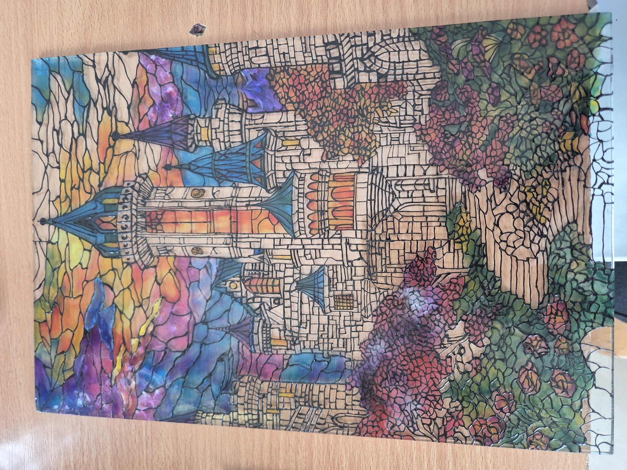 A colorful, stained-glass-style drawing on a clear surface, likely glass or acrylic, depicts a fanciful castle scene. The castle, centrally located, features multiple towers with blue-hued roofs and brick-like textures outlined in black. The central tower has a prominent orange and yellow facade. The architecture is a blend of fantasy and medieval inspiration.
The castle is surrounded by stylized landscapes. To the left, are colorful, abstract rock formations colored in shades of blue, purple, yellow, and orange. To the right of the castle are dense foliage and roses, rendered with red and green hues. The background is filled with a network of geometric shapes.
The entire drawing is placed on a light brown, wooden surface, which provides a simple backdrop. There is no evidence that this image is depicting any location in the city of Pavlikeni, Bulgaria. There are no people depicted in the drawing.