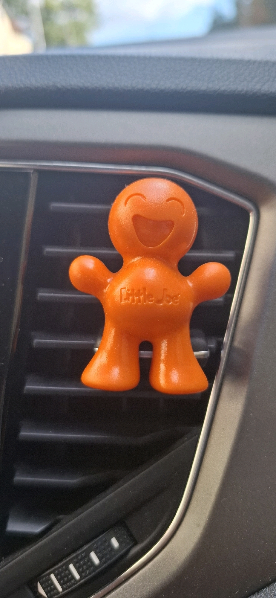An orange plastic car air freshener in the shape of a smiling gingerbread man is attached to a horizontal car air vent. The air freshener has "Little Joe" embossed on its chest. The air vent is black and has horizontal slats. A textured grey control dial is visible below the air vent, and a chrome-colored trim piece surrounds the vent. The background is blurred and appears to be the interior of a car, with a hint of an outdoor scene visible through a window. The image is a close-up shot, focusing on the air freshener and its immediate surroundings. The location is not specified in the image itself, and therefore cannot be determined.
