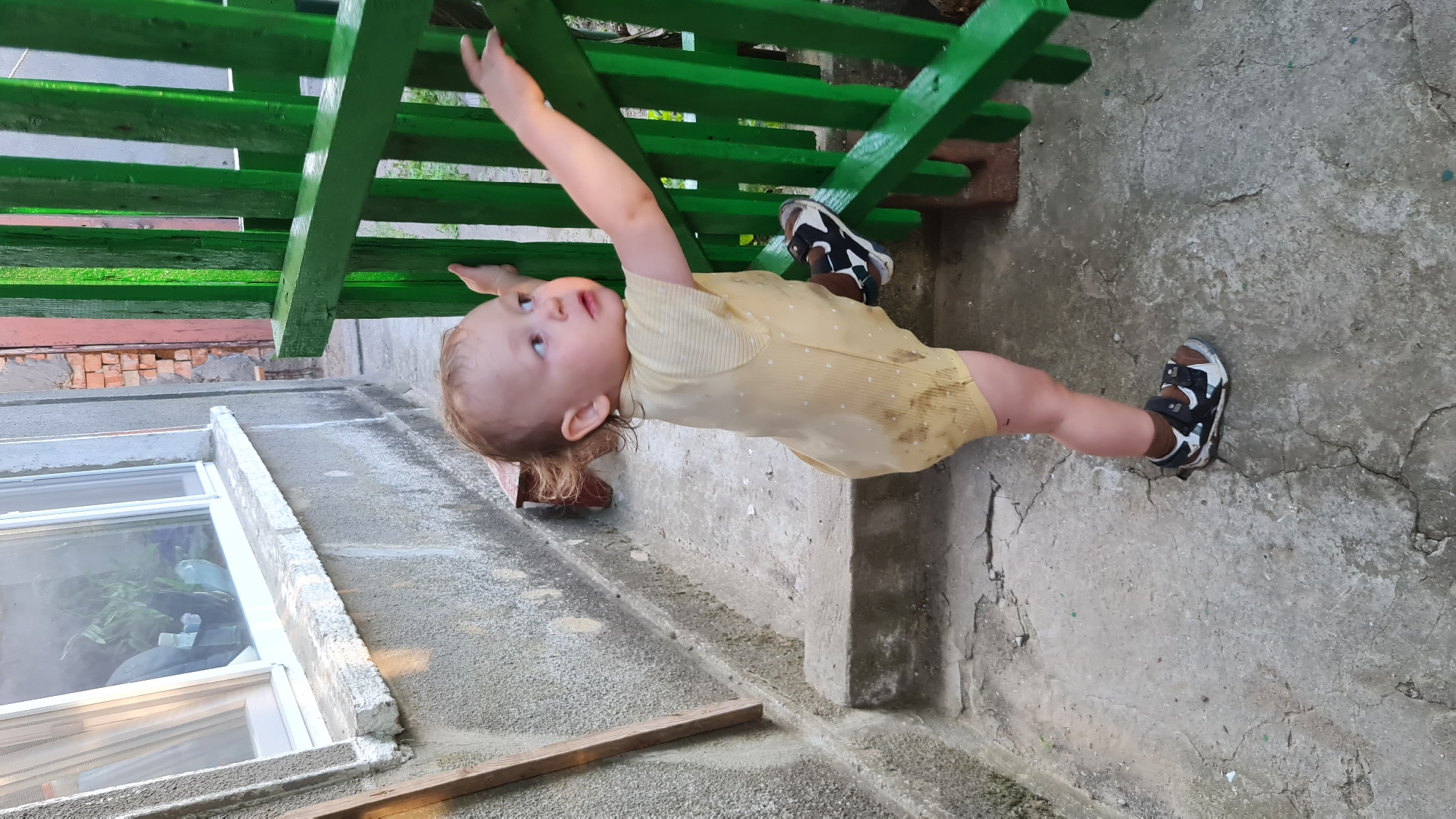 A young child, appearing to be around one to two years old, stands on a cracked concrete surface next to a set of outdoor stairs. The child is wearing a light yellow romper with small white dots and dark socks with sandals. They are reaching up towards a bright green wooden fence, with their right foot propped on the lower railing. The background shows a concrete wall with a window, some brickwork, and more of the green fence. The scene appears to be an outdoor area adjacent to a building, possibly in a residential setting within the city of Salhany, Ukraine. The overall impression is one of a candid moment of a child exploring their surroundings.
